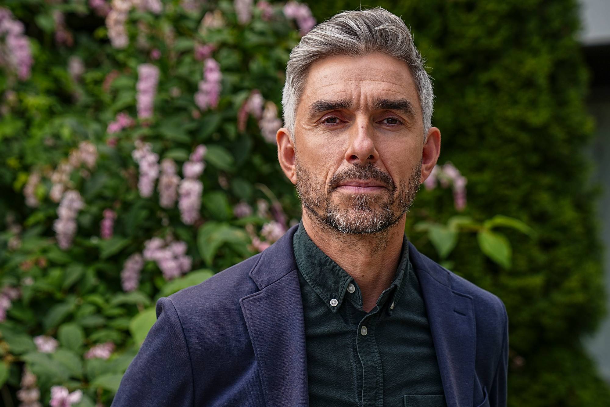 Et bilde av en mann med gråsprengt hår og skjegg. Han er kledd i en mørkeblå blazer og en mørk skjorte. Bak mannen er det busker og trær. (Bildebeskrivelsen er laget av en KI-tjeneste)