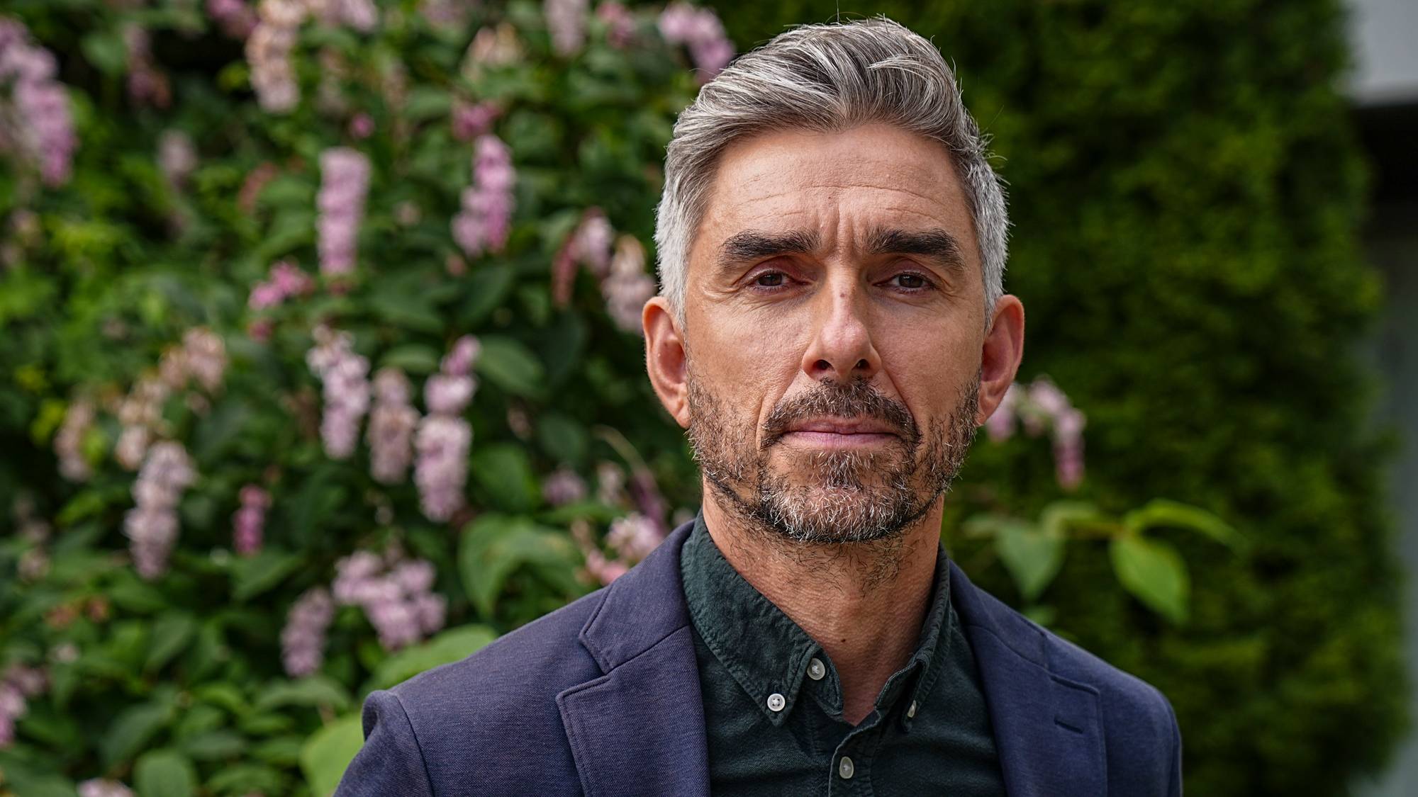 Et bilde av en mann med gråsprengt hår og skjegg. Han er kledd i en mørkeblå blazer og en mørk skjorte. Bak mannen er det busker og trær. (Bildebeskrivelsen er laget av en KI-tjeneste)