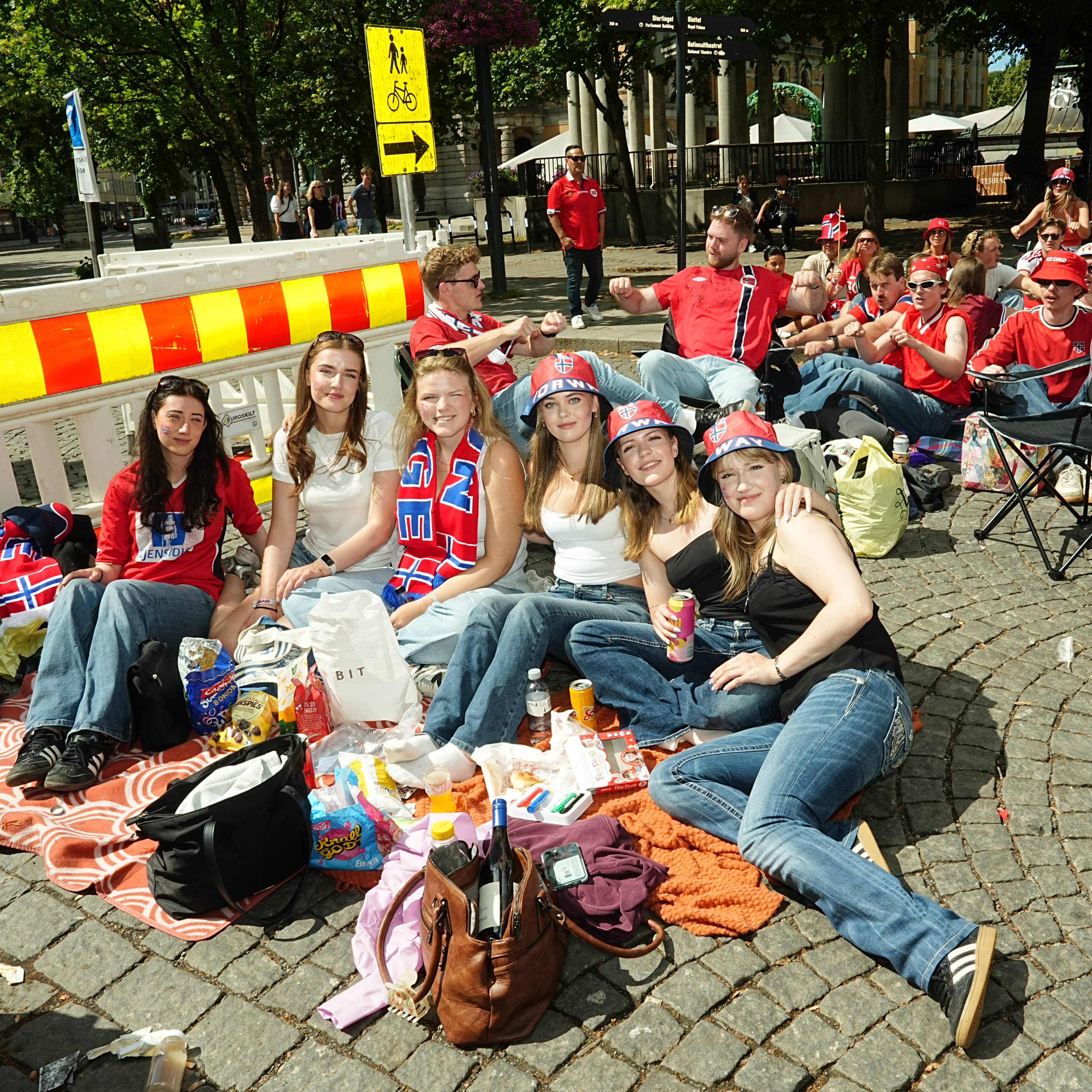Folk sitter på et teppe og stoler utendørs i en bygate under en markering eller folkefest. (Bildebeskrivelsen er laget av en KI-tjeneste)