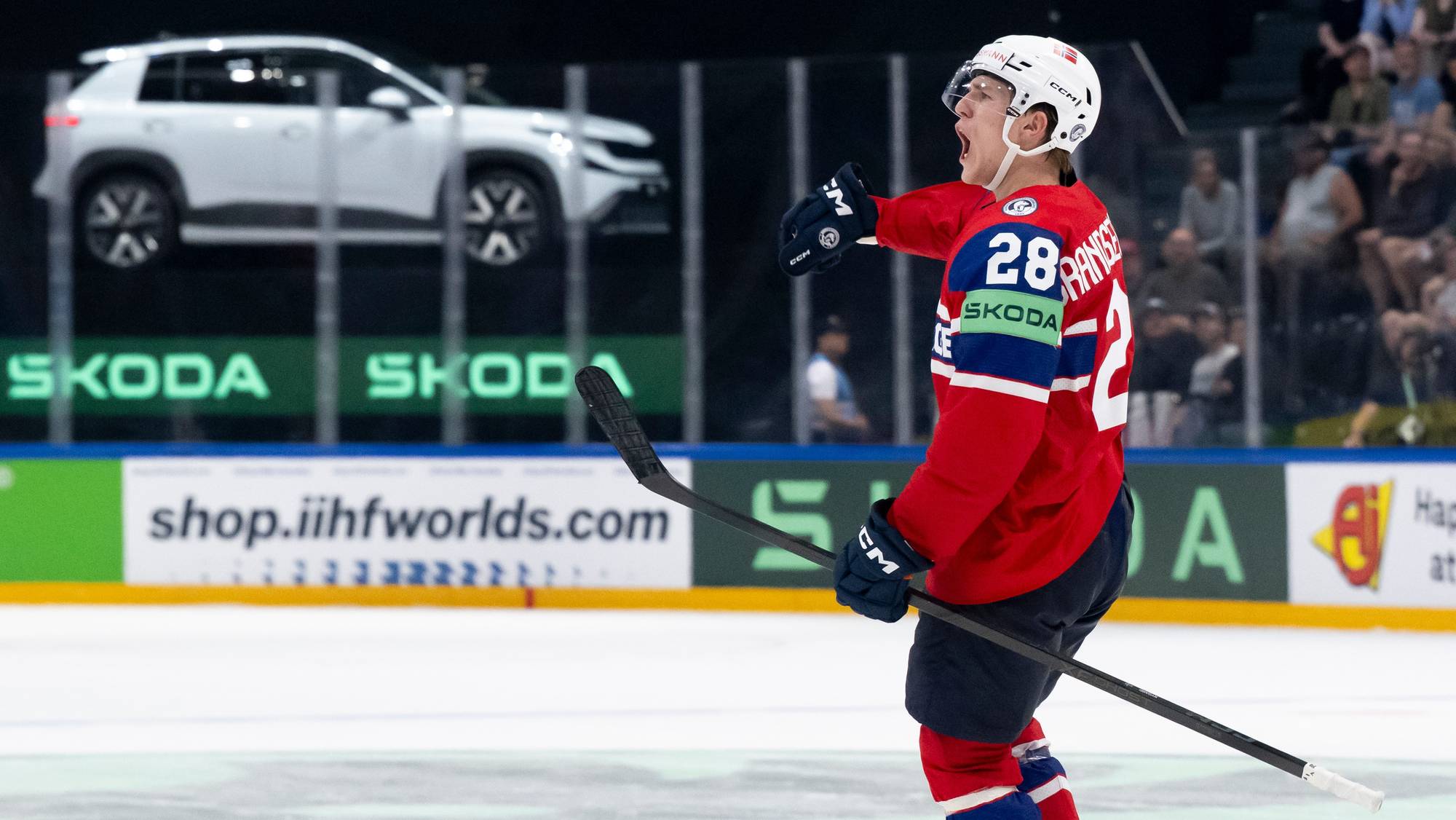 En ishockeyspiller i rød drakt med nummer 28 jubler. I bakgrunnen er det en hvit bil som henger i luften. Bak bilen er det grønne reklamebannere med "SKODA" på. (Bildebeskrivelsen er laget av en KI-tjeneste)