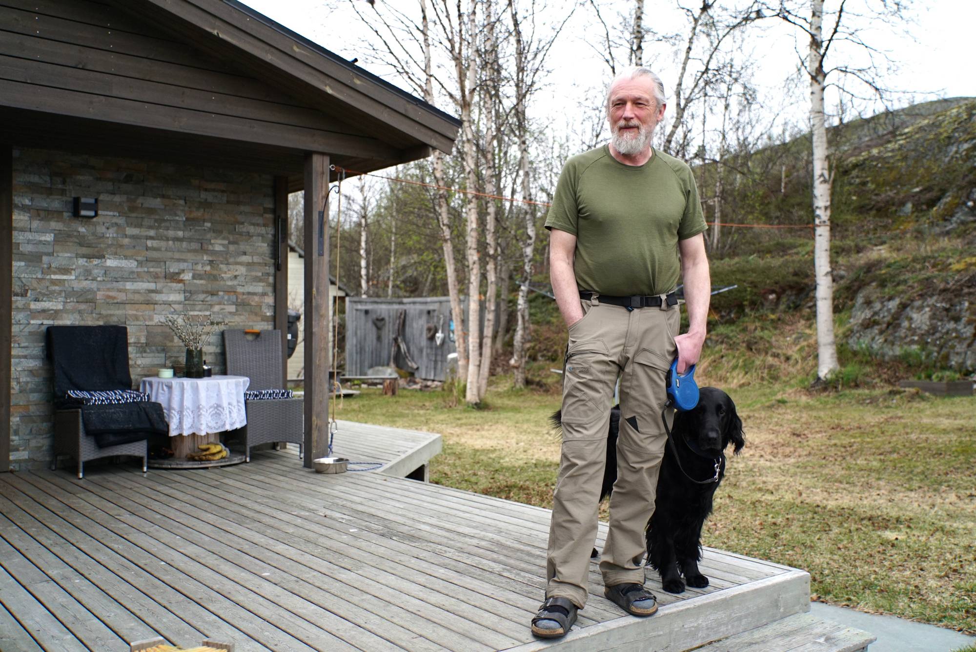 En mann med grått hår i hestehale med grønn t-skjorte og beige bukser som står ute på en trapp og holder en sort hund i bånd