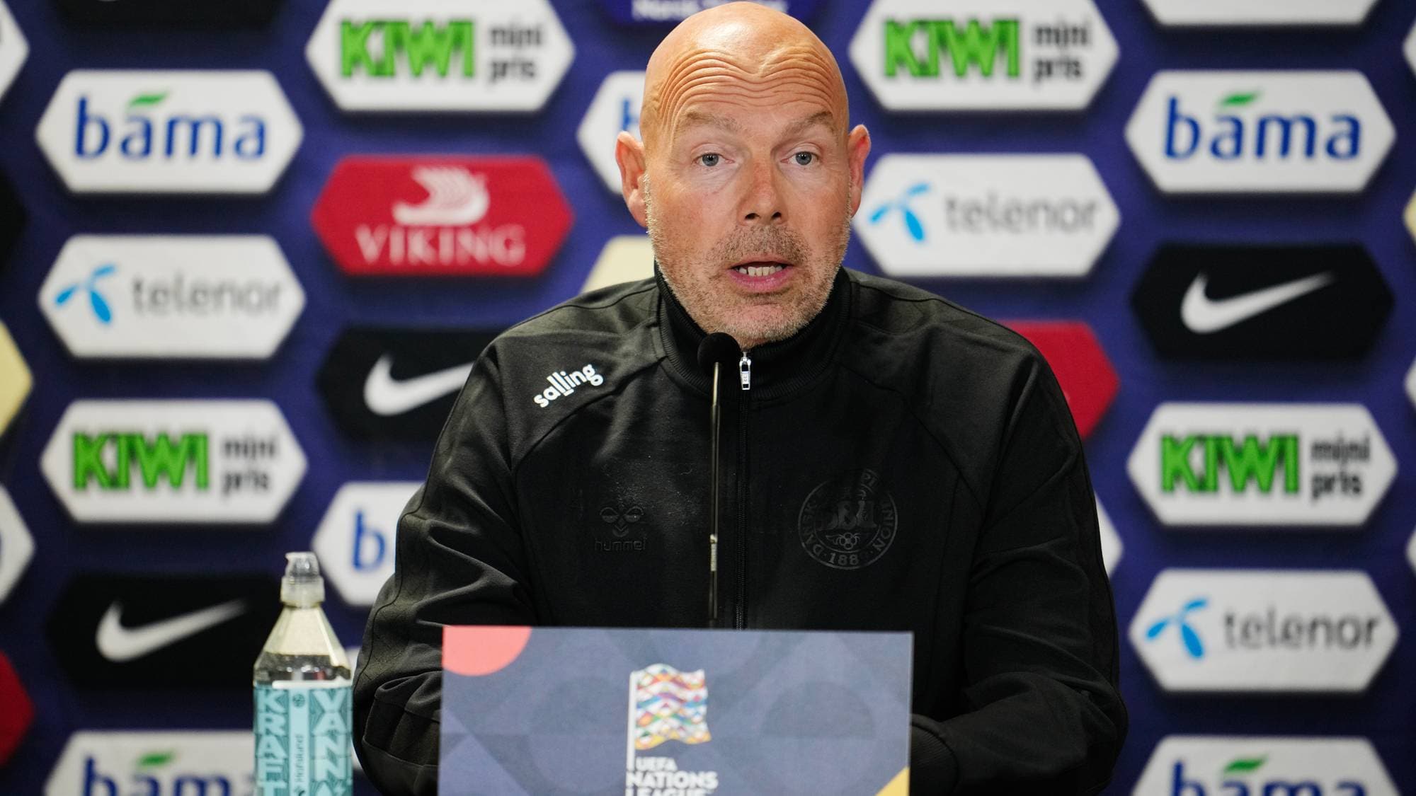 Danmarks landslagstrener Brian Riemer snakker i mikrofon under en pressekonferanse. (Bildebeskrivelsen er laget av en KI-tjeneste)