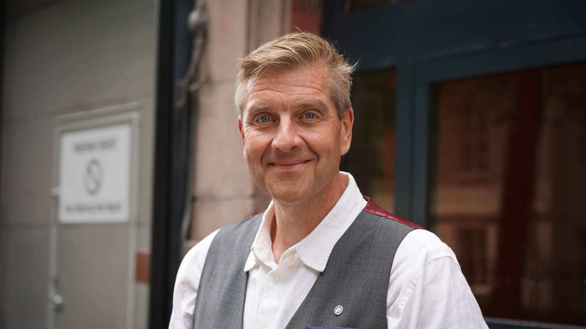 Portrett av en smilende mann i hvit skjorte og grå vest utendørs. 