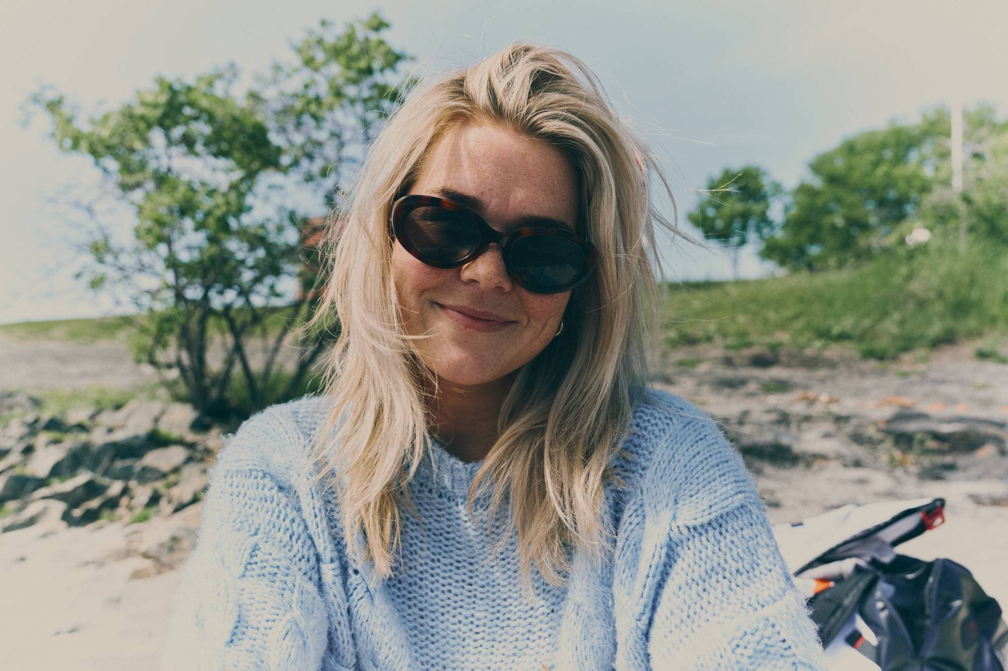Ung kvinne med sobriller og blondt hår smiler inn i kamera. 