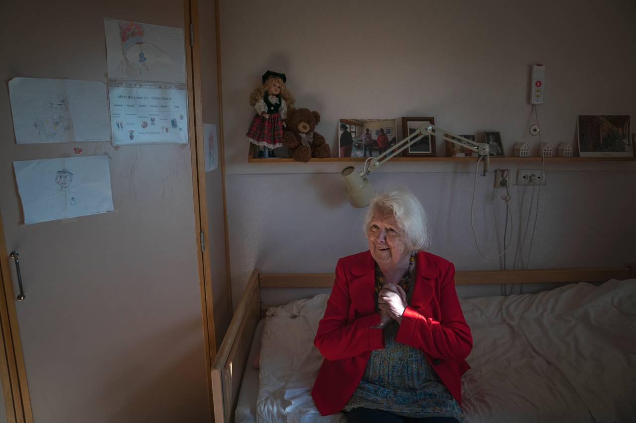 Kvinne i rød jakke sitter i seng og knytter hendene mens hun smiler stort. På veggen bak ser man en hylle med en rekke fotografier på en, en dukke, en teddybjørn og noen barnelignende tegninger på veggen ved siden av.