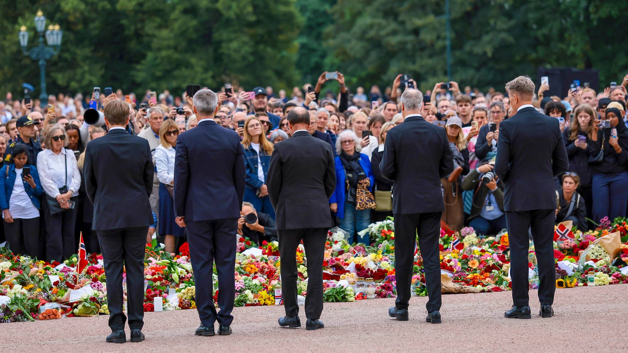 Fem regjeringsmedlemmer, blant annet Jens Stoltenberg og Espen Barth Eide, sett bakfra. Foran dem et blomsterhav og et folkehav på Slottsplassen.