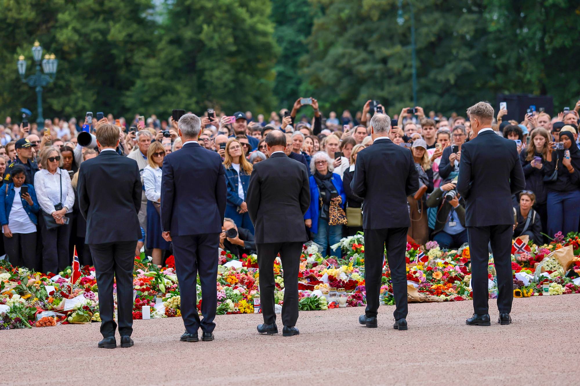 Fem regjeringsmedlemmer, blant annet Jens Stoltenberg og Espen Barth Eide, sett bakfra. Foran dem et blomsterhav og et folkehav på Slottsplassen.