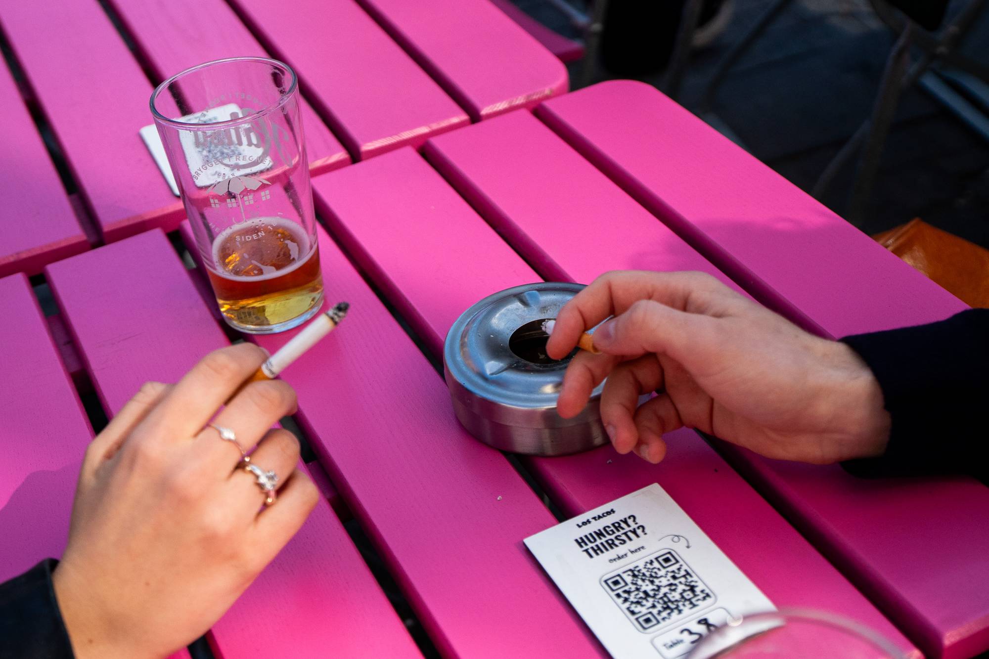 Et glass øl og en sigarett ligger på et rosa trebord. En person holder en brennende sigarett med høyre hånd og en annen person holder en sigarett med filteret vendt mot et askebeger. Et lite papir med en QR-kode og teksten "HUNGRY? THIRSTY?" ligger på bordet. (Bildebeskrivelsen er laget av en KI-tjeneste)