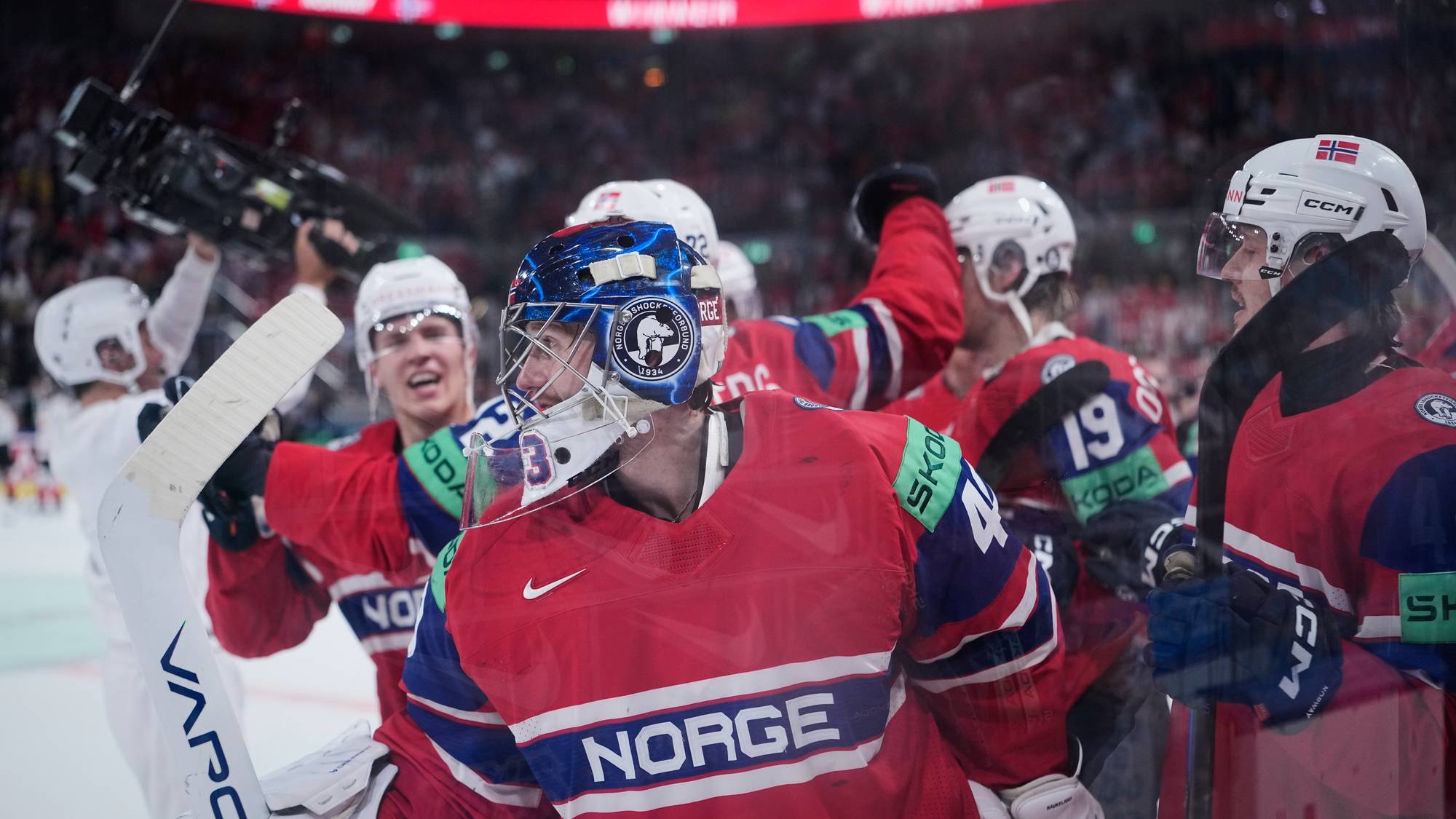 Ishockeylandslaget feirer seier. Spillere i røde og blå drakter med Norges flagg på hjelmen. En spiller med maske og kølle er i forgrunnen. (Bildebeskrivelsen er laget av en KI-tjeneste)
