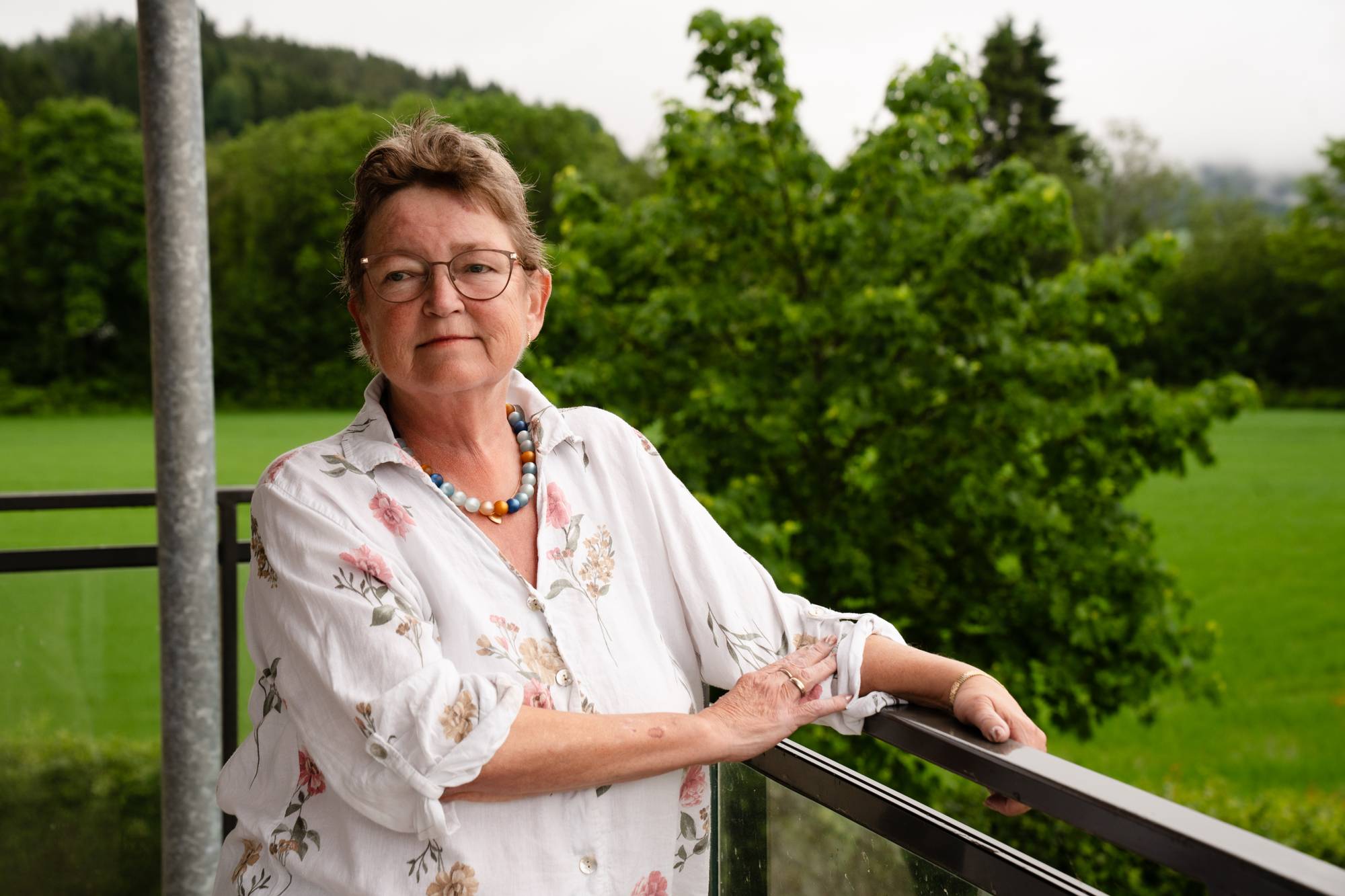 En eldre kvinne med briller og blomstrete bluse står på en balkong. Hun ser mot kameraet. Bak henne er det grønne trær og gress. En grå stolpe er synlig til venstre i bildet. (Bildebeskrivelsen er laget av en KI-tjeneste)