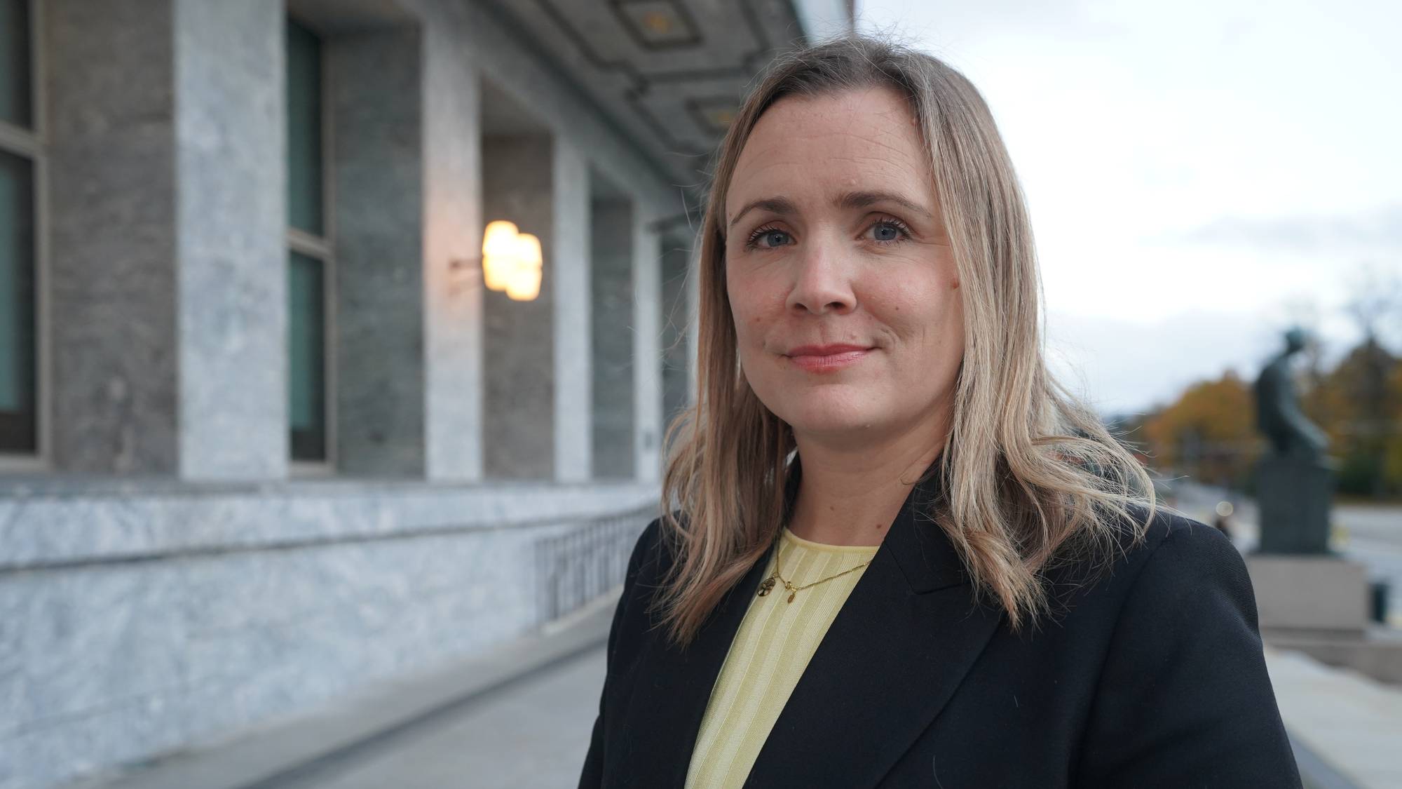 Marit Vea, en kvinne med langt, lyst hår står ved rådhuset i Oslo, iført en mørk jakke over en lys gul genser