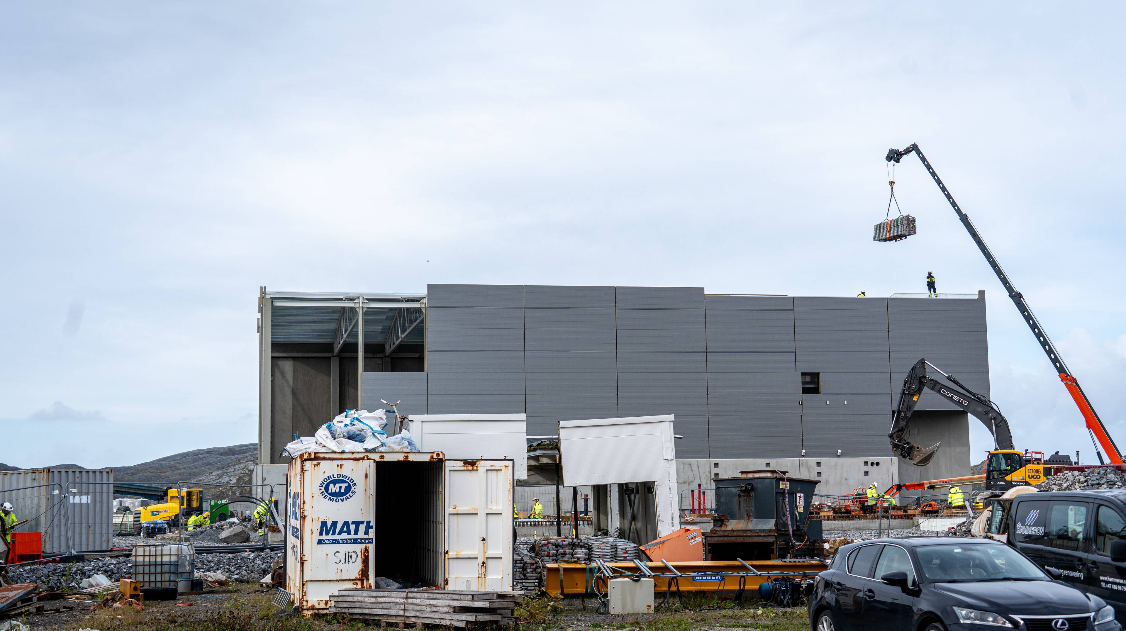 Bildet viser en byggeplass, byggematerialer og en kran som heiser materialer på et tak, mens en arbeider tar i mot. Det skal bli en del av den nye hydrogenfabrikken ved innseilinga til Bodø, som Green H bygger for å produsere hydrogen til de nye hydrogenfergene til Lofoten.