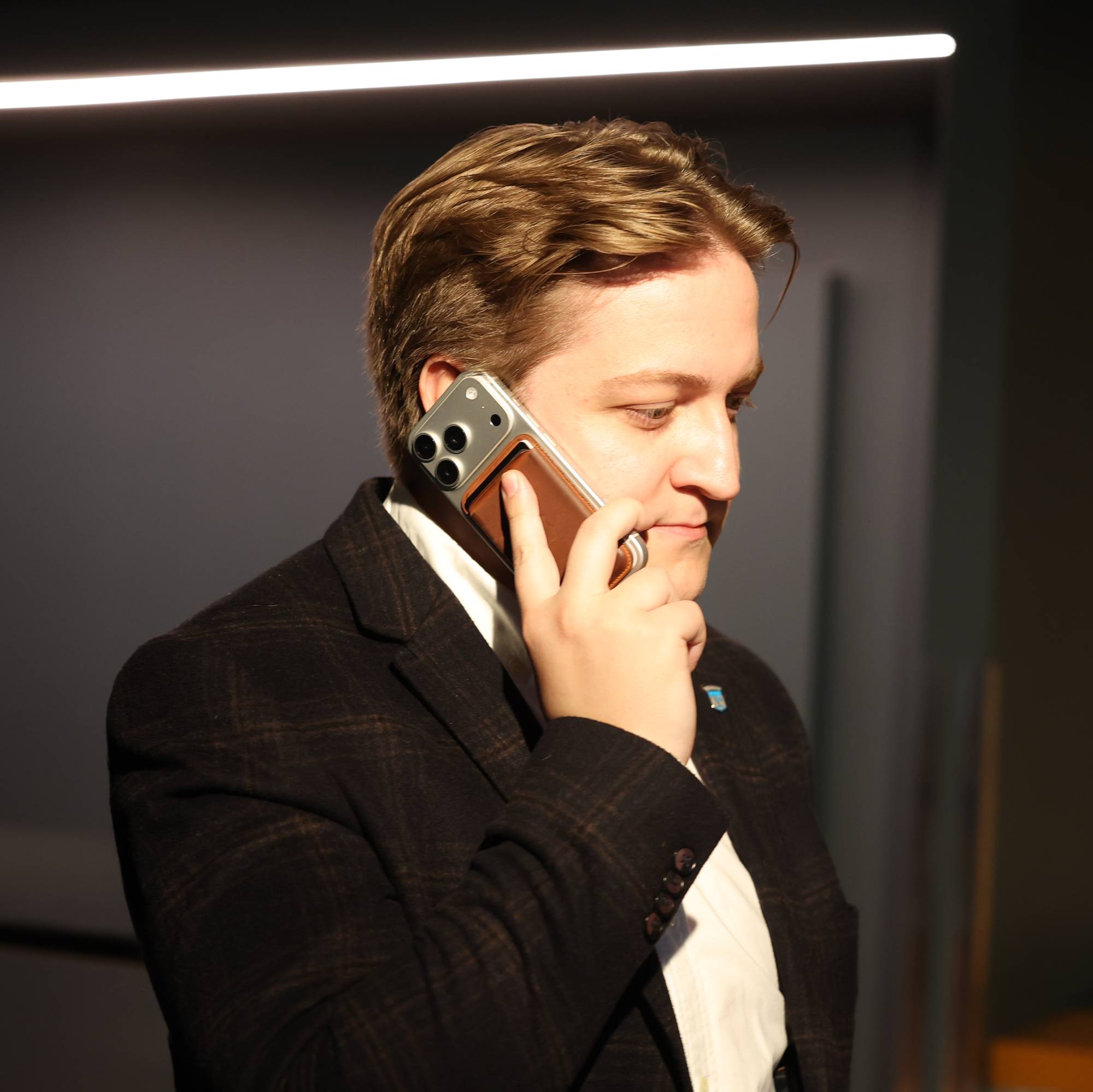 En mann med brunt ruteformet dressjakke og hvit skjorte snakker i en mobiltelefon. Han holder telefonen til høyre øre med høyre hånd. Mannen ser ned. Bak ham er en vegg og en lysstripe i taket. (Bildebeskrivelsen er laget av en KI-tjeneste)