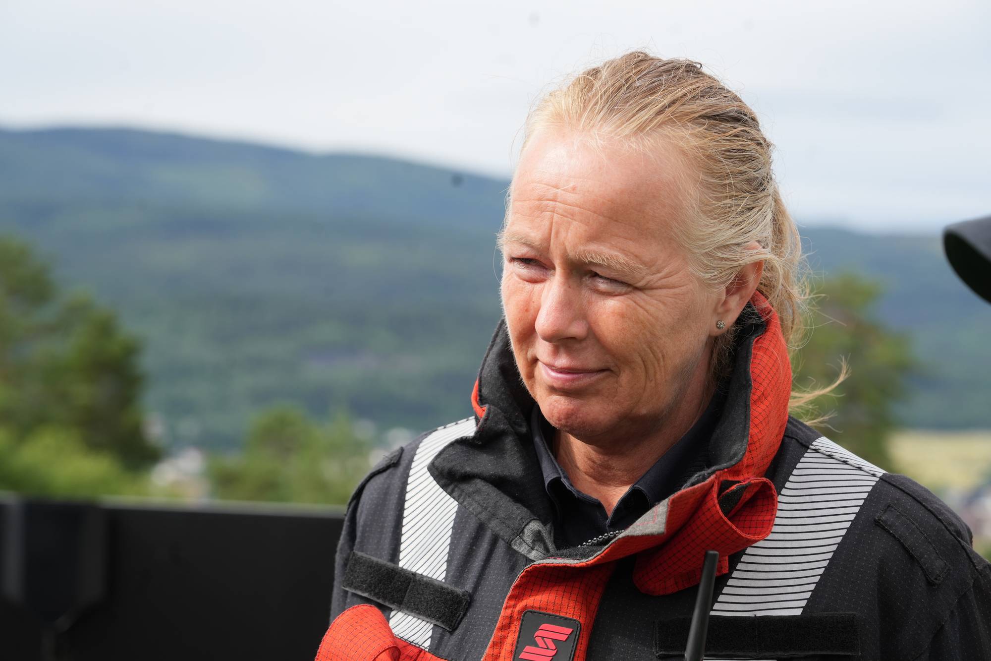 Kvinne med lyst hår i hestehale kledd i rød og svart uniform med refleksdetaljer. Ute i landskap. (Bildebeskrivelsen er laget av en KI-tjeneste)
