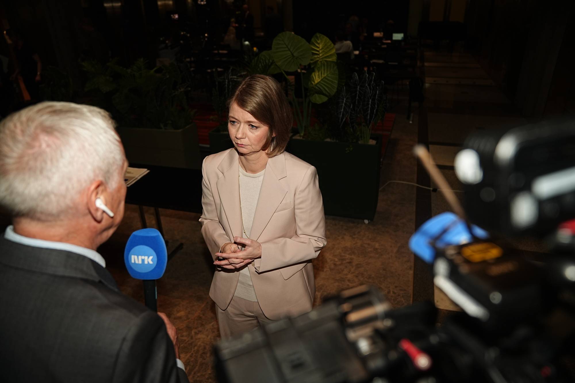 En kvinne i lys dress blir intervjuet foran en NRK-mikrofon. En reporter står foran henne med ryggen til kameraet. I forgrunnen er et TV-kamera. Bak dem er grønne planter og mørke omgivelser. (Bildebeskrivelsen er laget av en KI-tjeneste)