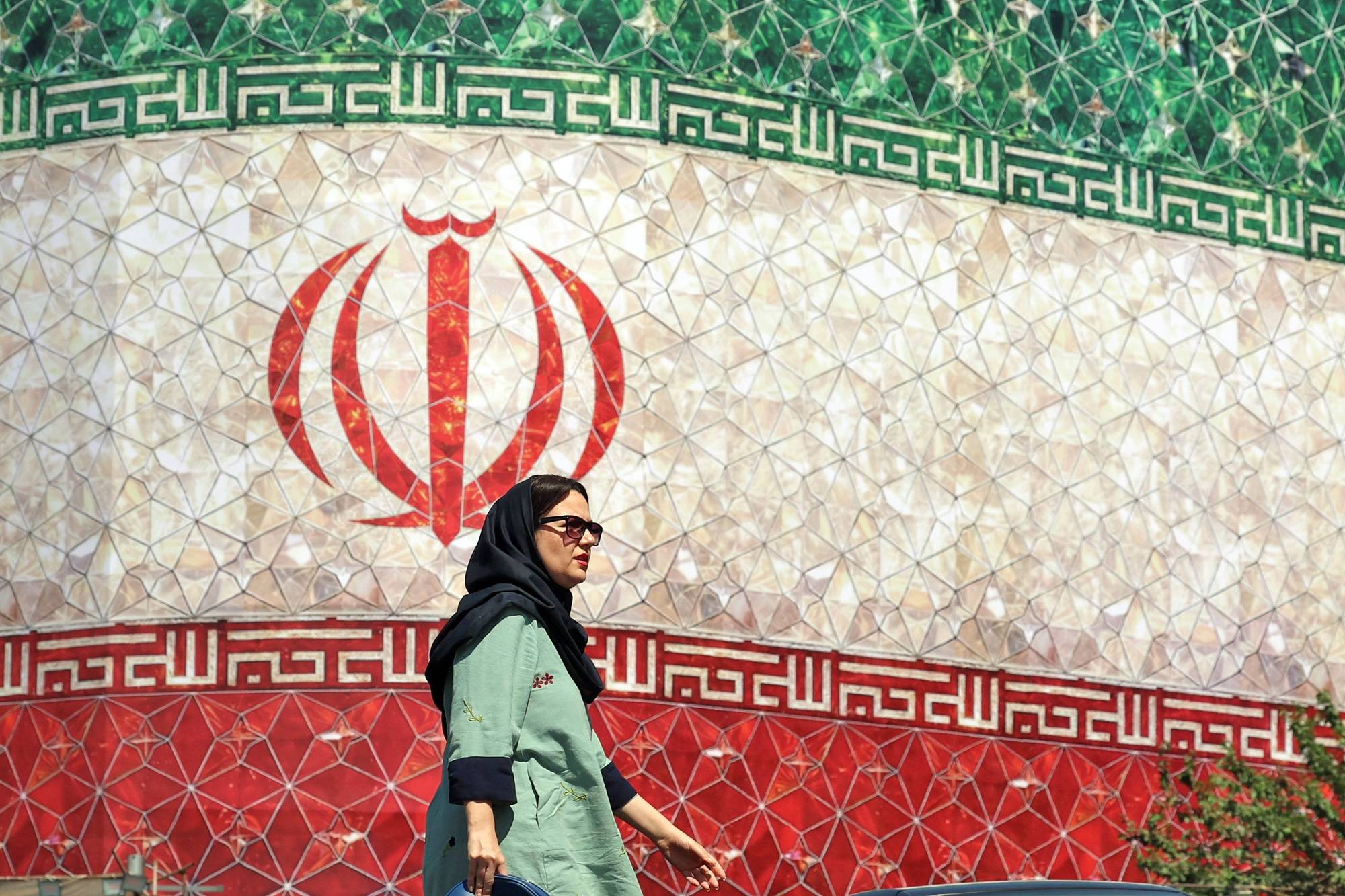 En kvinne med hijab og solbriller går forbi et stort, mosaikkbelagt bilde av Irans flagg. Mosaikken dekker en stor bygningsfasade. Bildet er tatt utendørs i dagslys. (Bildebeskrivelsen er laget av en KI-tjeneste)