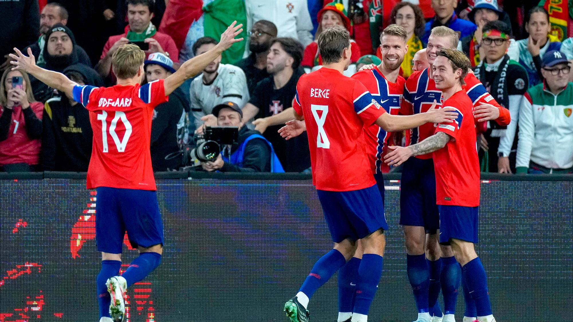 Norske fotballspillere i røde drakter feirer mål sammen på banen foran publikum. (Bildebeskrivelsen er laget av en KI-tjeneste)