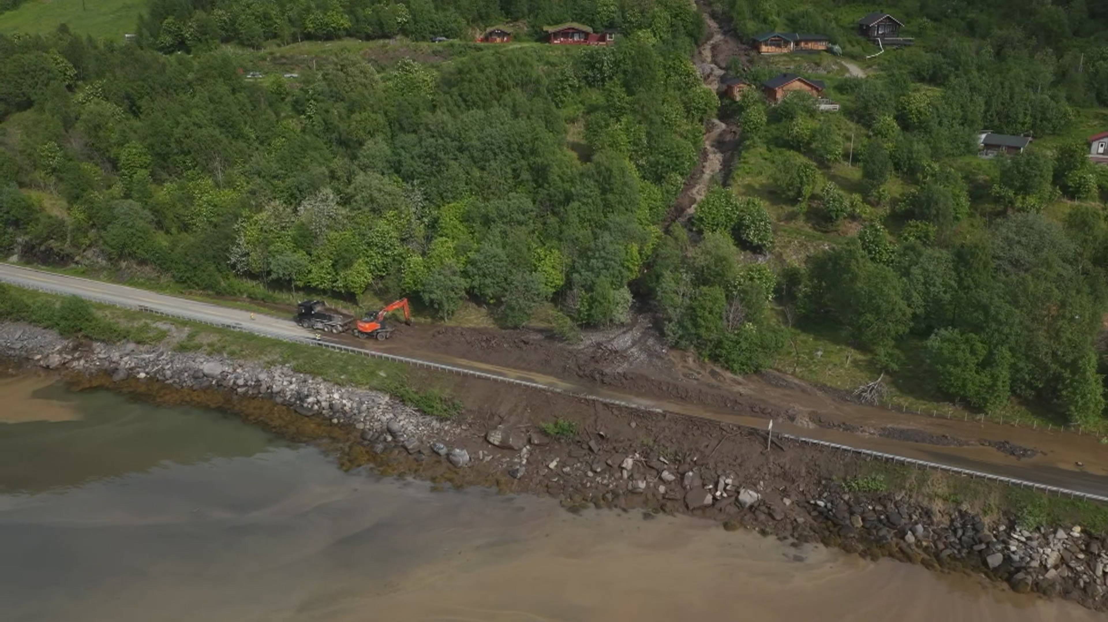 Et anleggskjøretøy og en gravemaskin jobber ved en vei som er delvis dekket av jord. Jorden ser ut til å ha rast ned fra en skråning hvor flere hus ligger. Veien grenser til et vann. (Bildebeskrivelsen er laget av en KI-tjeneste)