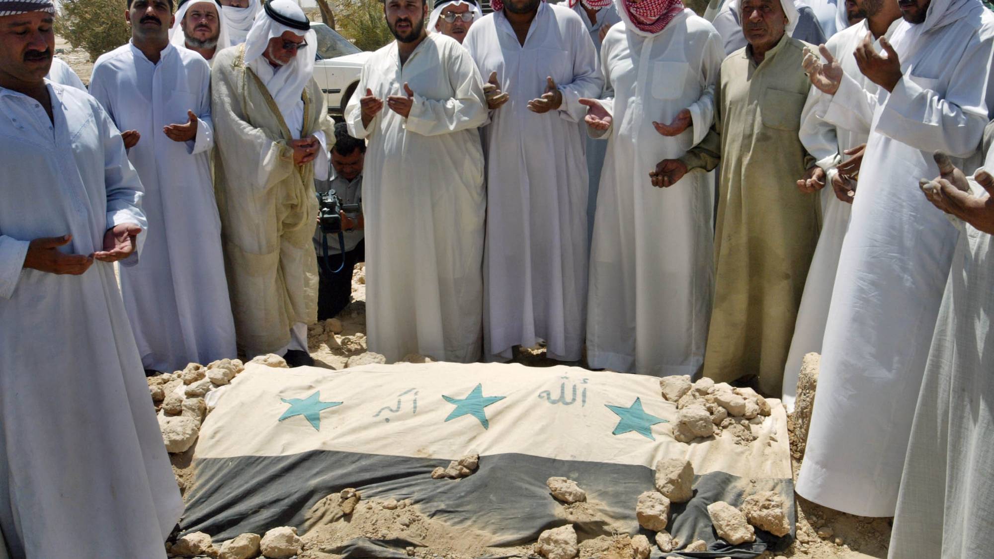 En gruppe menn i tradisjonelle klær samlet rundt et likdekt av et flagg. Mennene holder hendene løftet i bønn. Liket er dekket av det irakiske flagget fra før 2004, som har tre grønne stjerner og arabisk skrift. Bakgrunnen er utydelig, men viser noen trær og en bil. (Bildebeskrivelsen er laget av en KI-tjeneste)