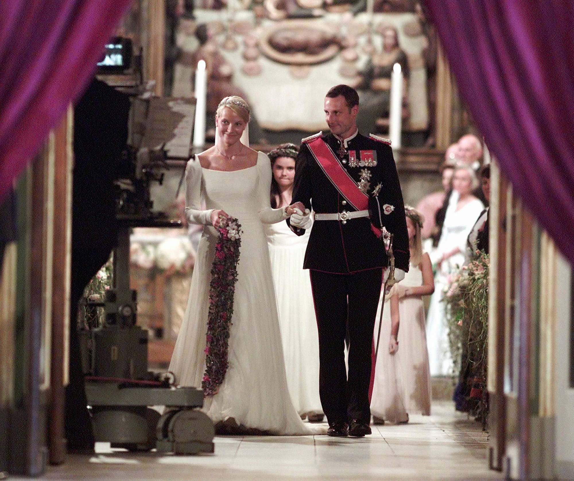 Kronprins Haakon og kronprinsesse Mette-Marit går arm i arm ut av Oslo domkirke etter å ha blitt viet 25. august 2001. Foran dem er et TV-kamera, og bak dem går brudepikene og brudesvennen.