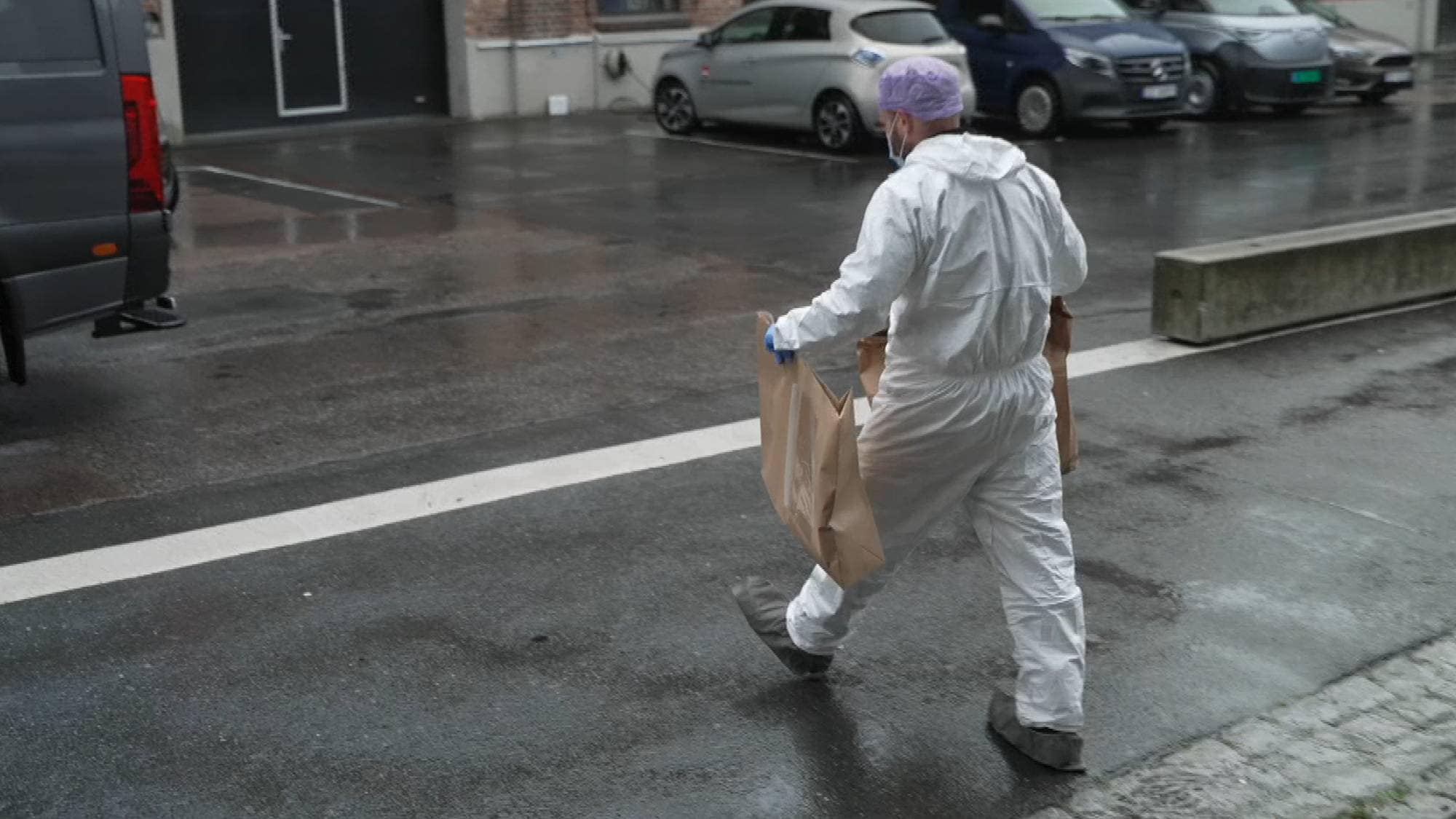 Person i hvitt beskyttelsesutstyr bærer papirposer over en våt parkeringsplass. (Bildebeskrivelsen er laget av en KI-tjeneste)