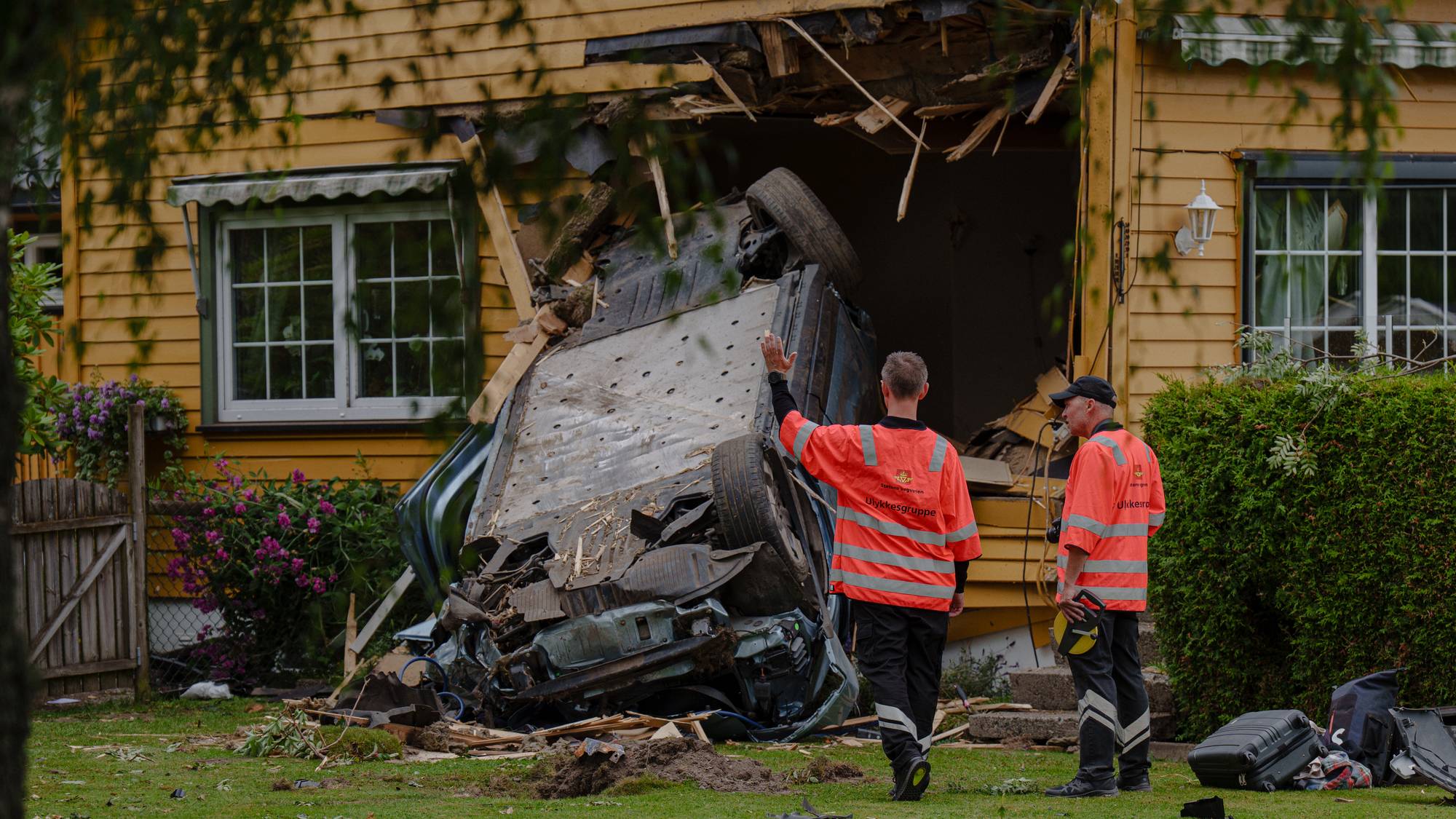 Bil har krasjet inn i en trevegg på bolighus. To personer i refleksvester undersøker skadestedet. (Bildebeskrivelsen er laget av en KI-tjeneste)