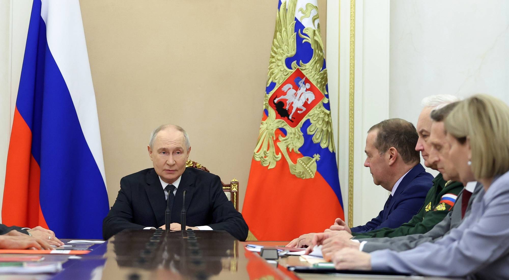 Presidnet Vladimir Putin sitter ved det tradisjonelle bordet sitt. mellom to russiske flagg. Ved han sitter andre russiske politkere, men han er i sentrum. Han har en seriøs mine, og rynker i pannen. 