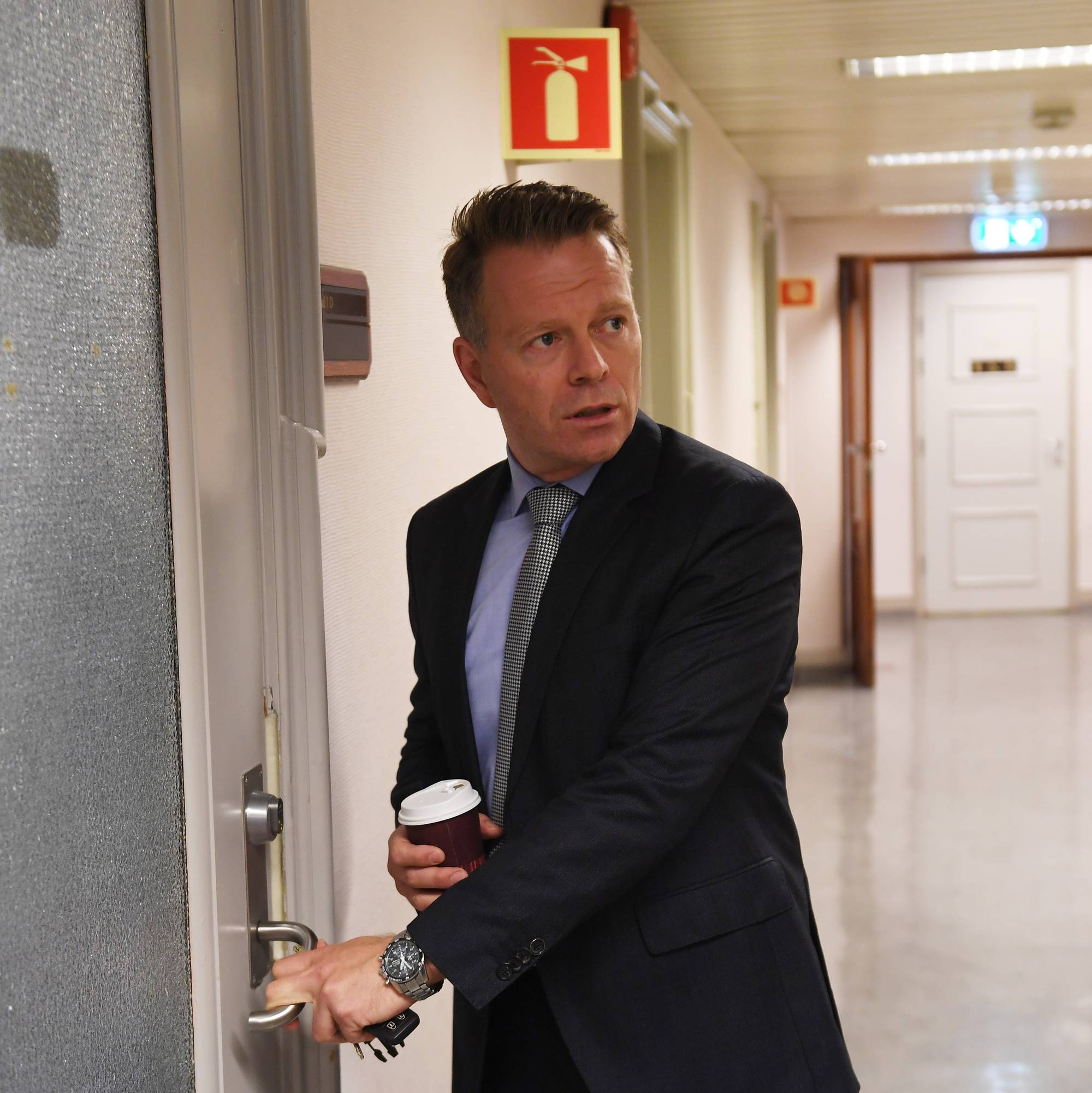 Mann i dress med kaffekopp åpner en dør i en kontorkorridor. (Bildebeskrivelsen er laget av en KI-tjeneste)