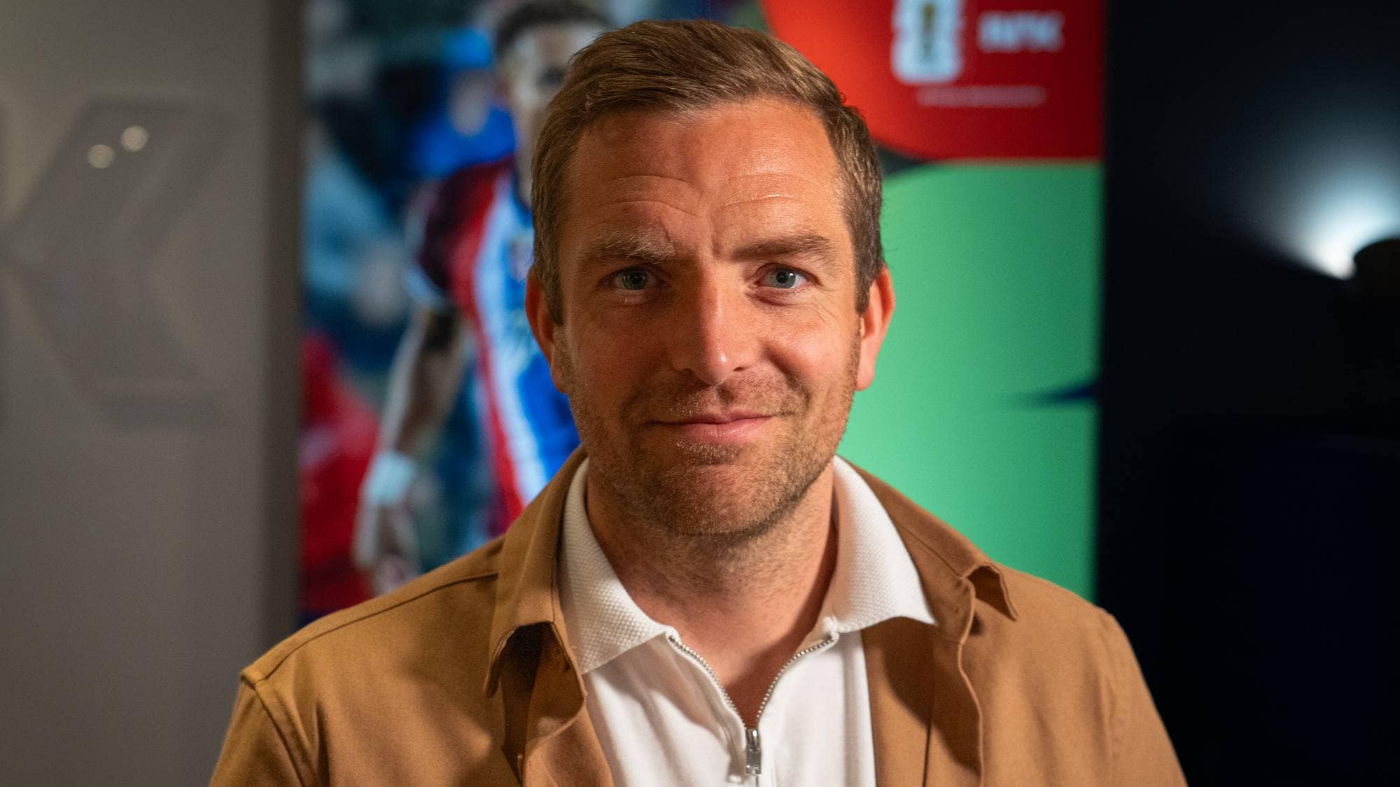 Mann i brun jakke og hvit kragegenser ser mot kamera. Bak ham er et bilde av en fotballspiller og en grønn bakgrunn med en logo. Han har kort, lyst hår og blå øyne. Bildet er tatt på nært hold. (Bildebeskrivelsen er laget av en KI-tjeneste)
