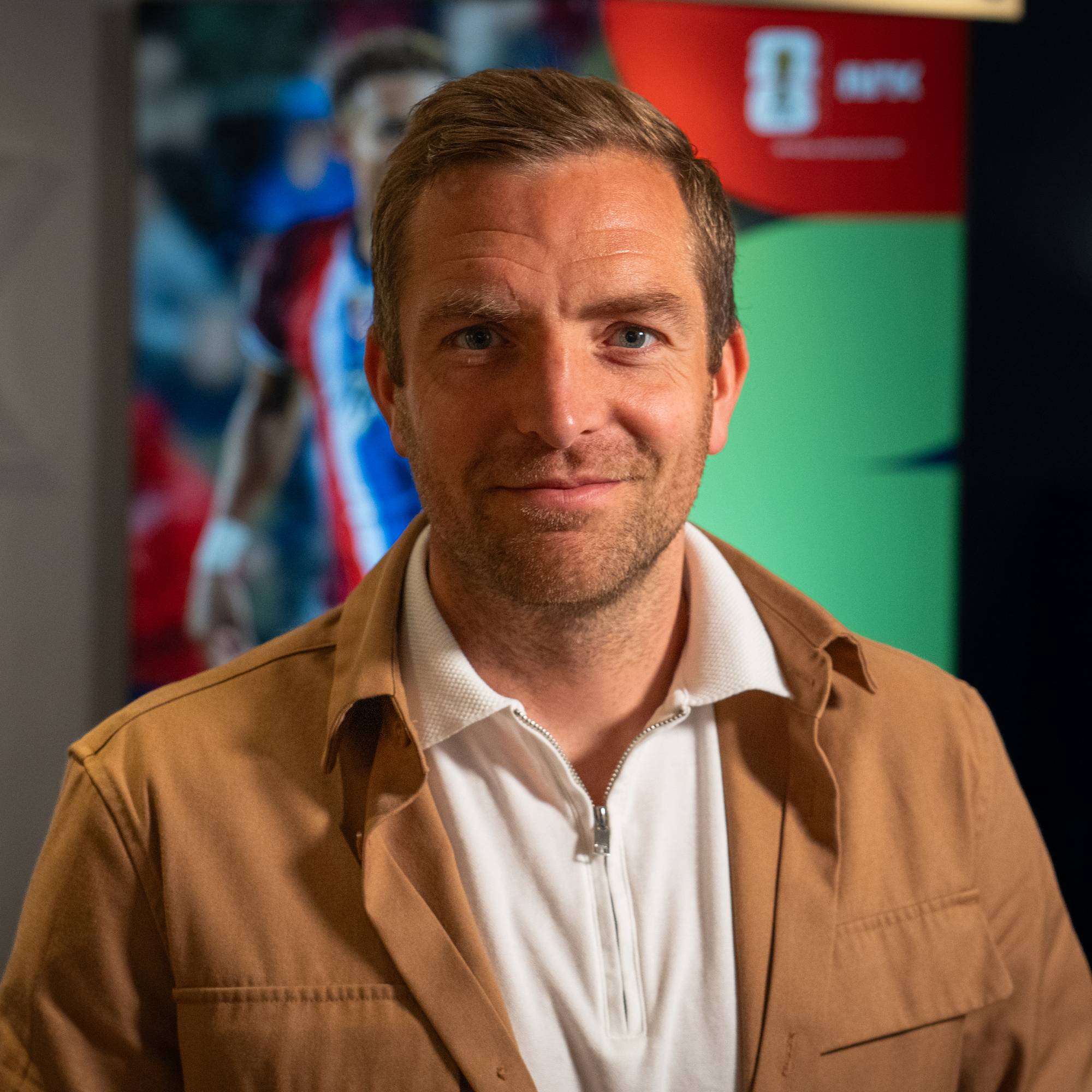 Mann i brun jakke og hvit kragegenser ser mot kamera. Bak ham er et bilde av en fotballspiller og en grønn bakgrunn med en logo. Han har kort, lyst hår og blå øyne. Bildet er tatt på nært hold. (Bildebeskrivelsen er laget av en KI-tjeneste)