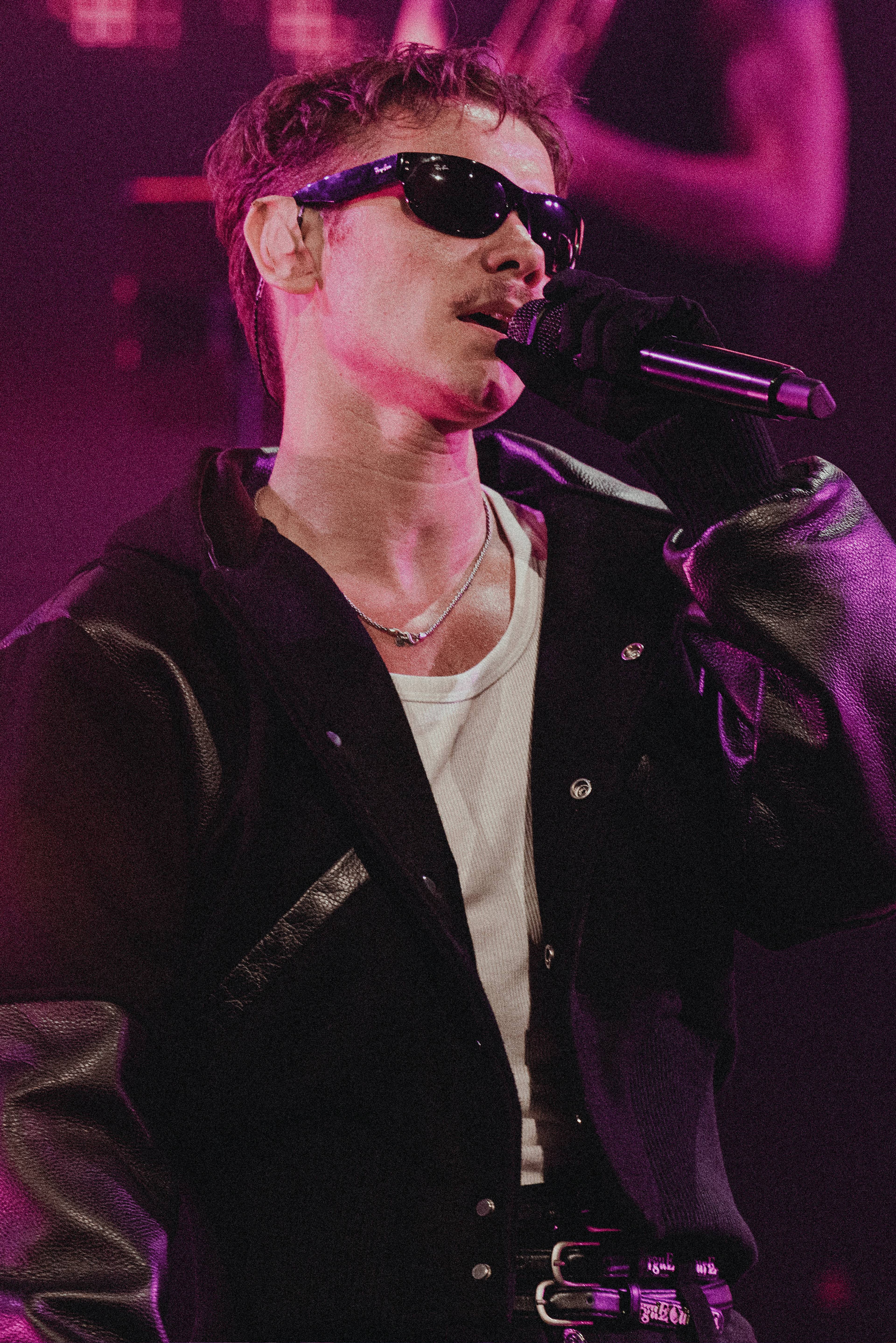 Artist Carl Louis ser i solbriller mens han synger i mikrofon. Han er kledd i en svart jakke med hvite detaljer. Bildet er tatt på en konsert med lilla scenebelysning. (Bildebeskrivelsen er laget av en KI-tjeneste)