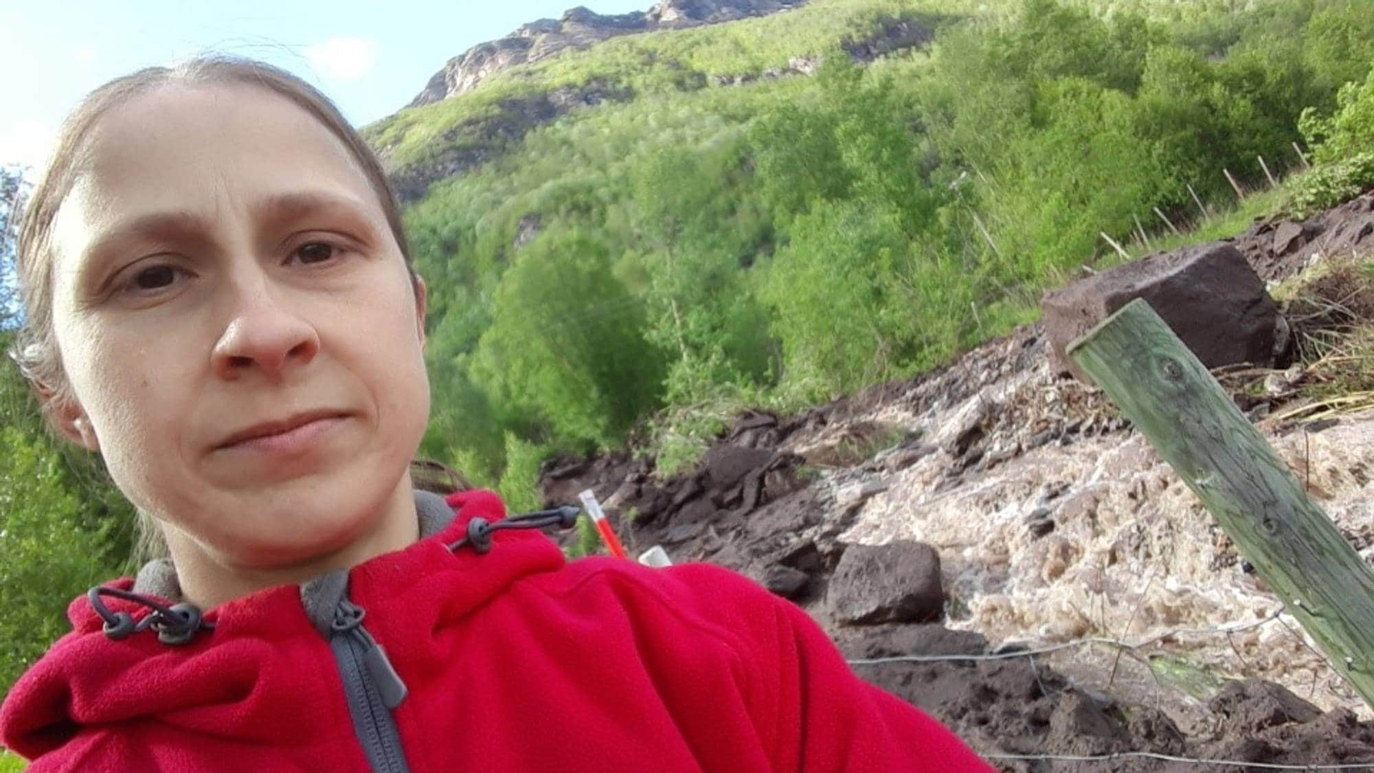 En kvinne i rød jakke ser mot kameraet. Bak henne er et fjell med grønn vegetasjon og spor etter et ras. (Bildebeskrivelsen er laget av en KI-tjeneste)