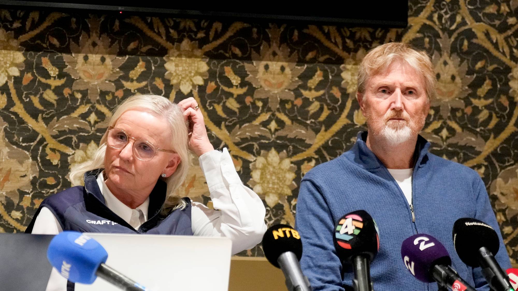 Tre personer sitter rundt et bord dekket med en hvit duk. På bordet er det en laptop, mikrofoner og en vannflaske. Veggen i bakgrunnen har et dekorativt mønster. Det ser ut som de forbereder seg til en presentasjon eller pressekonferanse. (Bildebeskrivelsen er laget av en KI-tjeneste)
