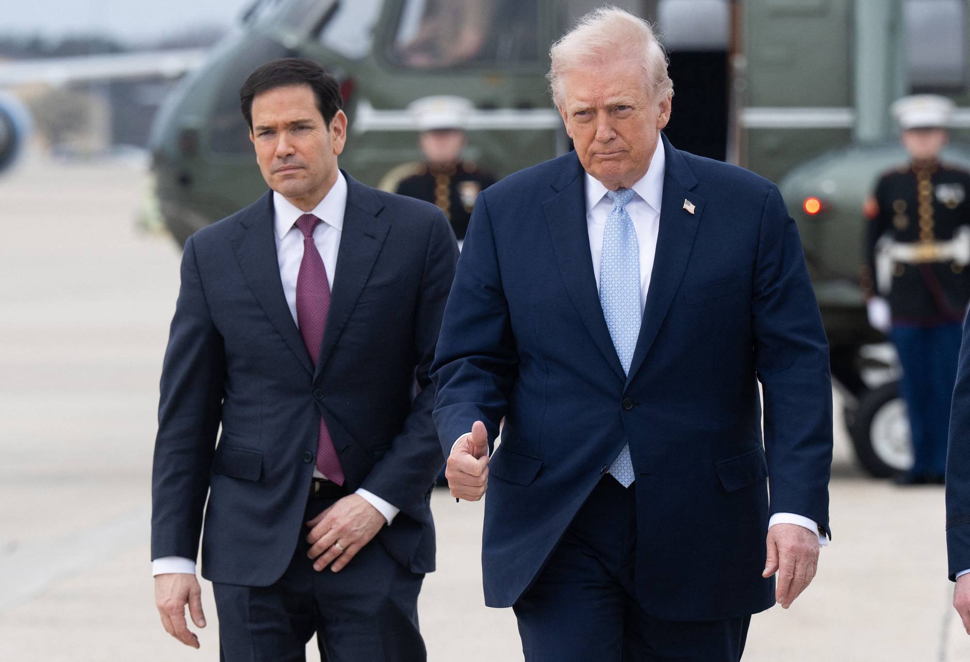 Donald Trump og Marco Rubio går ved siden av hverandre. Begge er kledd i dress og slips. Bak dem står det uniformerte personer. En helikopterlignende farkost er synlig i bakgrunnen. (Bildebeskrivelsen er laget av en KI-tjeneste)