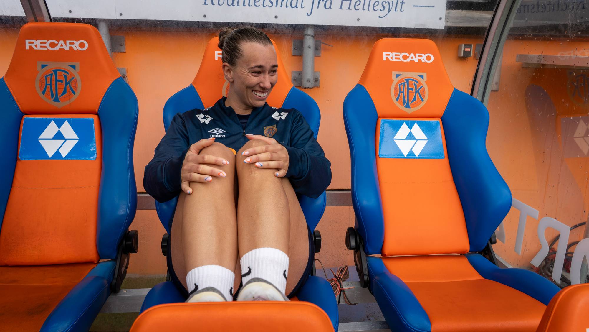 Smilende fotballspiller sitter på innbytterbenken med knærne trukket opp til brystet. (Bildebeskrivelsen er laget av en KI-tjeneste)