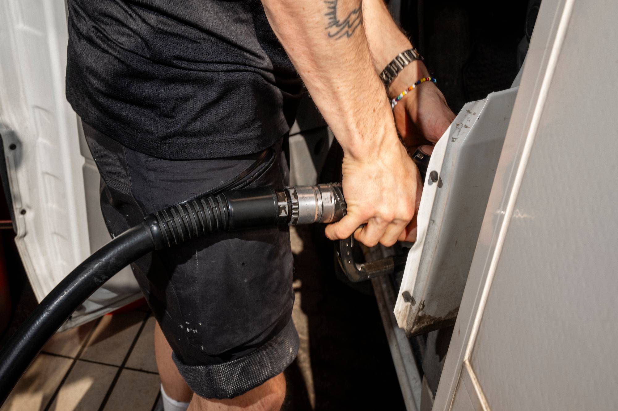 En person fyller drivstoff på et kjøretøy. Personen holder en drivstoffpumpe i hånden og setter den inn i påfyllingslokket. Han har på seg en svart t-skjorte og mørke shorts. Bilen er hvit. (Bildebeskrivelsen er laget av en KI-tjeneste)