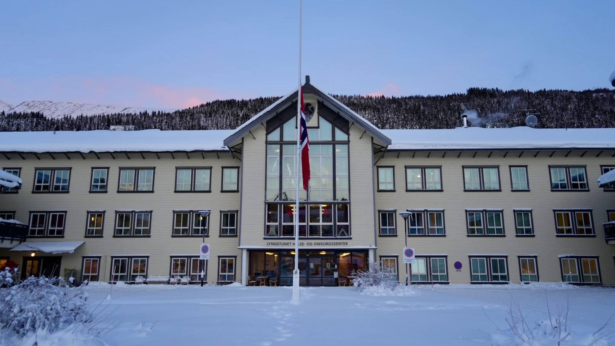 Bygningen er Lyngstunet helse- og omsorgssenter, dekket av snø. Flagget henger på halv stang foran inngangen. Det er en klar himmel med snødekte fjell i bakgrunnen. Omgivelsene er stille og vinterlige, med spor i snøen som tyder på aktivitet.