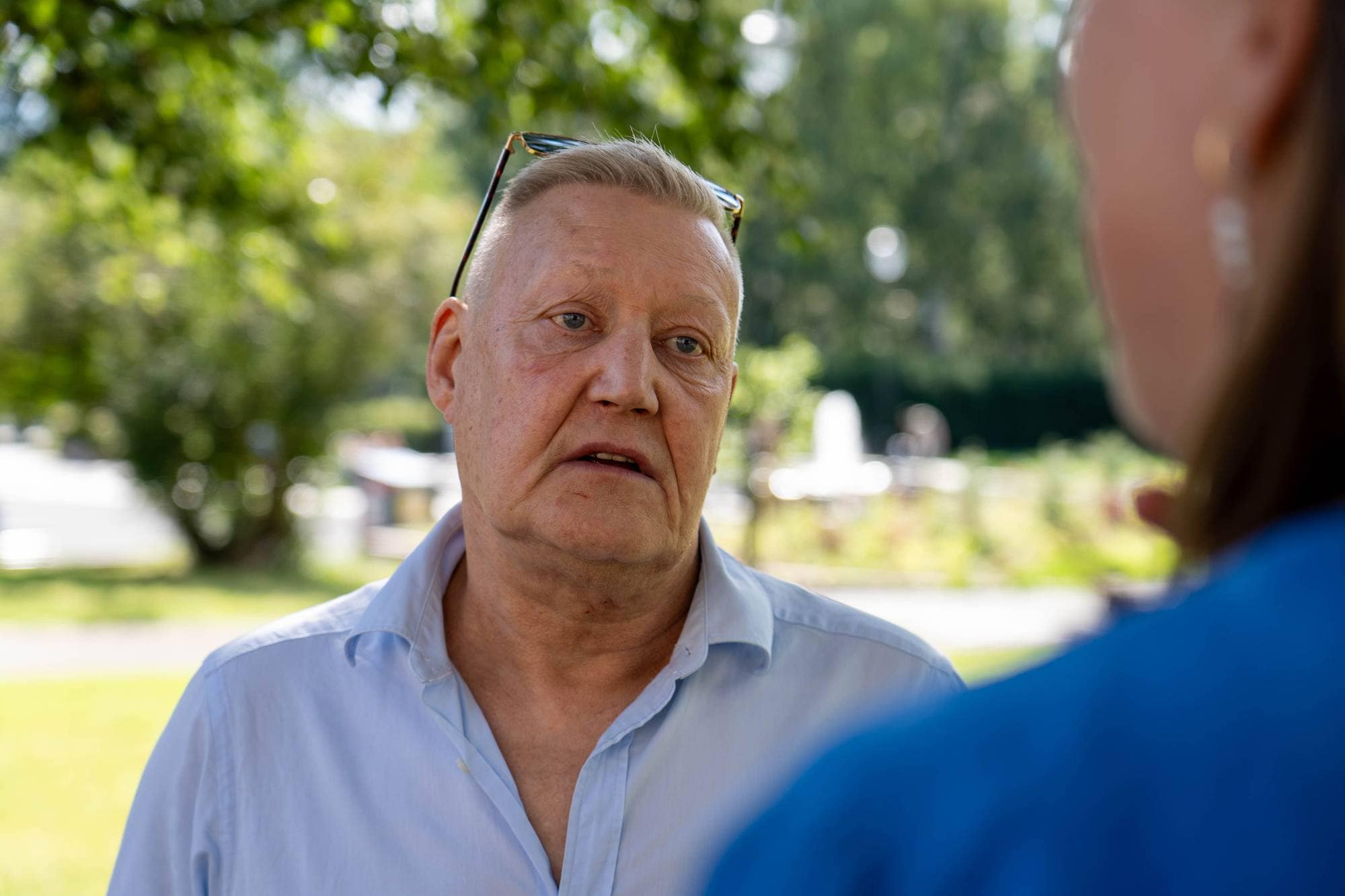 Eldre mann med briller i håret snakker med en person ut av bildet i en park. (Bildebeskrivelsen er laget av en KI-tjeneste)