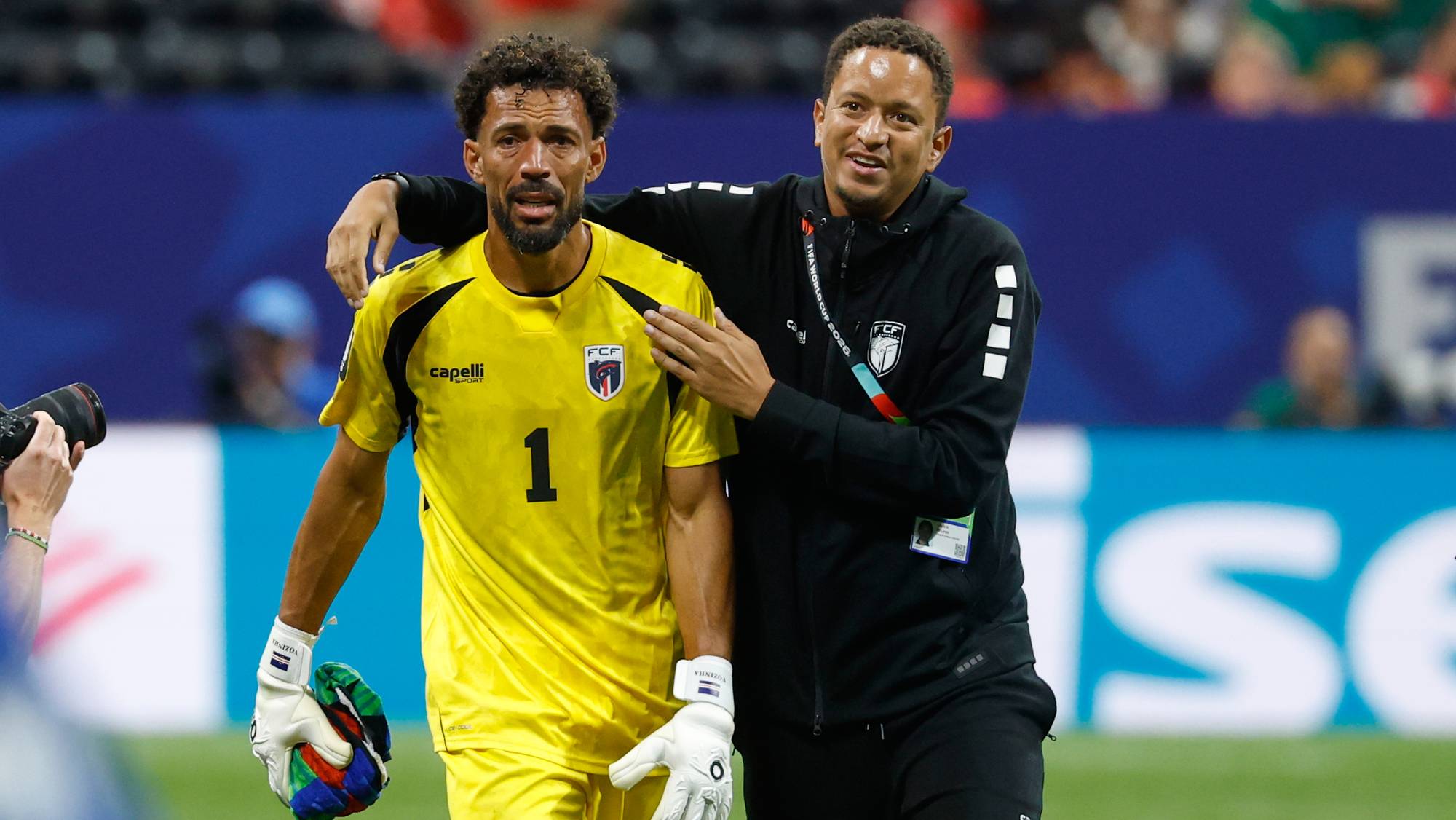 To fotballspillere er på banen. Den ene har på seg gul drakt og hansker, den andre har på seg svart treningsjakke. (Bildebeskrivelsen er laget av en KI-tjeneste)