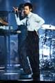 Scott A Garfitt / AP / NTB Harry Styles opptrer på scenen iført en lys skjorte og stripete bukser. Han holder en mikrofon og står med en dramatisk positur. I bakgrunnen ses musikere ved instrumenter og lyssetting som bidrar til en livlig atmosfære.