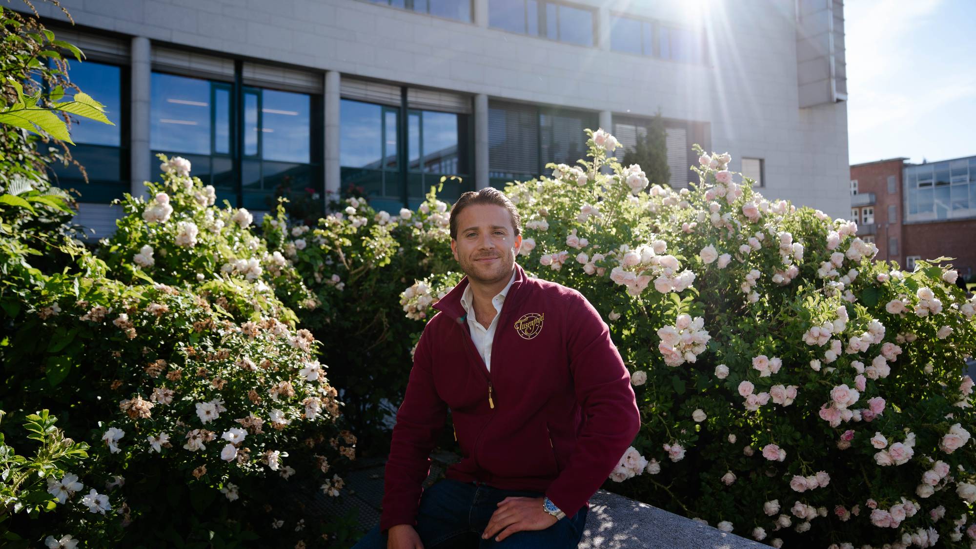 En mann sitter foran en busk med lyserosa blomster. Han er kledd i en vinrød genser og blå jeans. I bakgrunnen er en moderne bygning. 