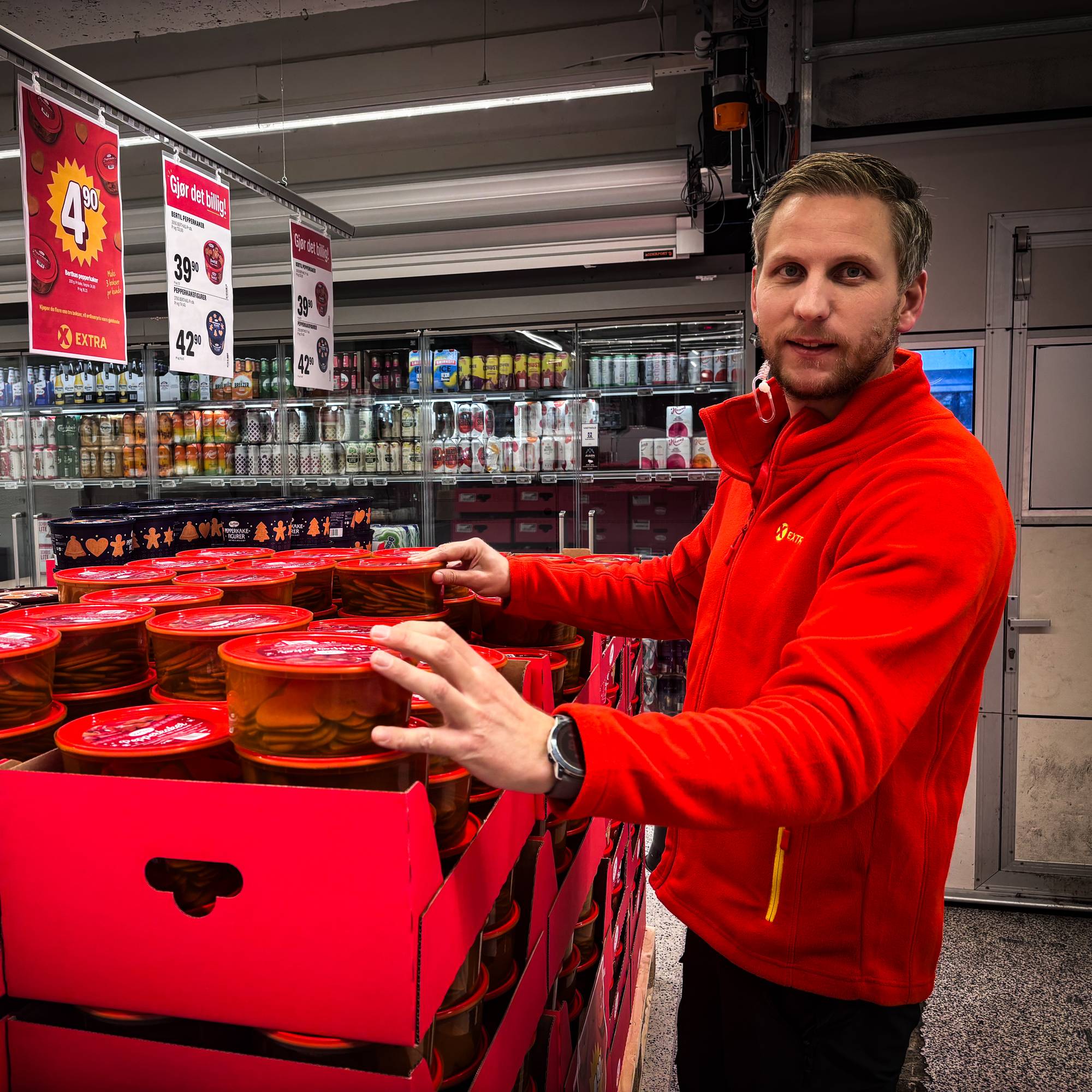 En mann i rød fleecejakke plasserer varer i en utstillingskasse i en dagligvarebutikk. (Bildebeskrivelsen er laget av en KI-tjeneste)