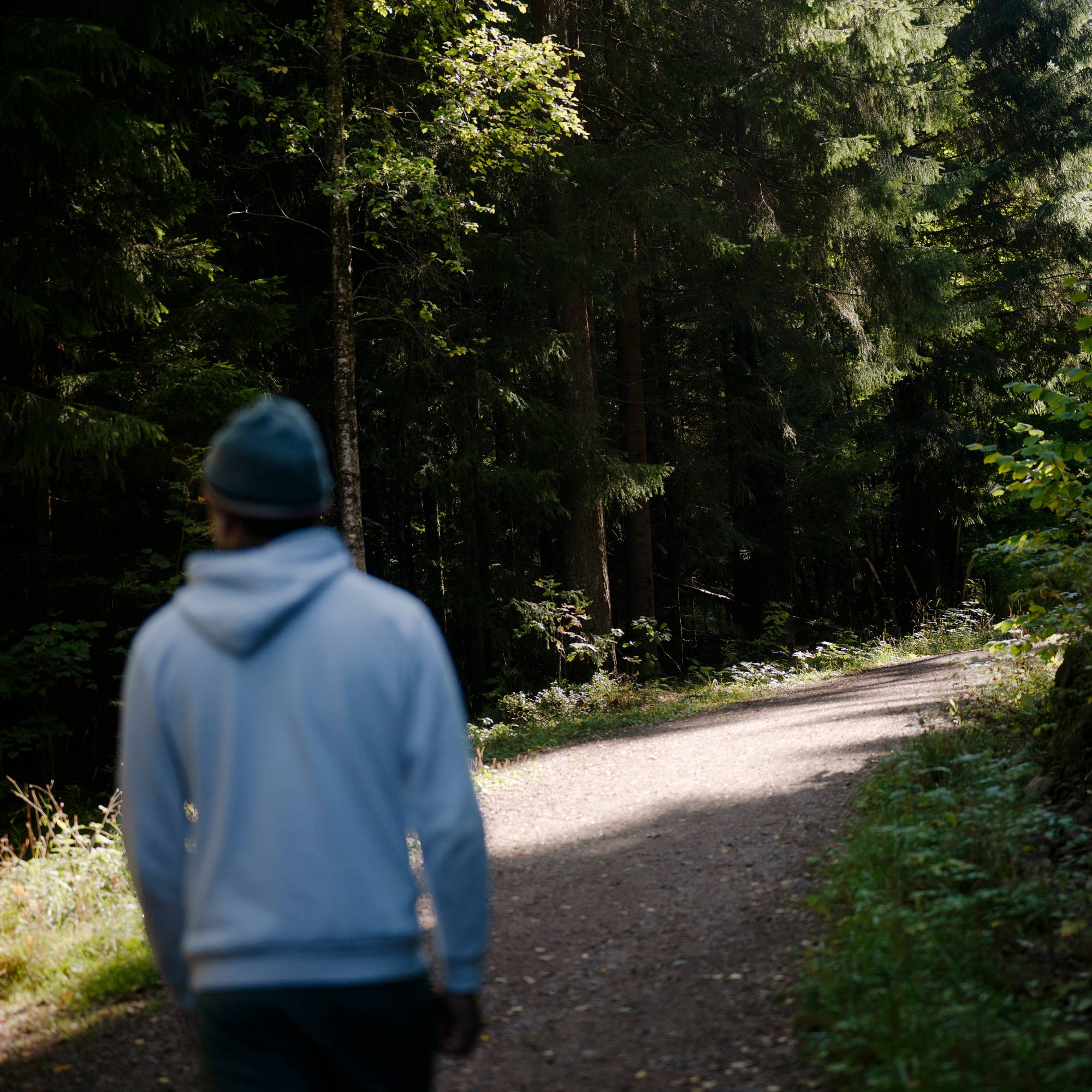 En gutt med hettegenser og lue går vekk fra kamera på en grusvei i en skog. 