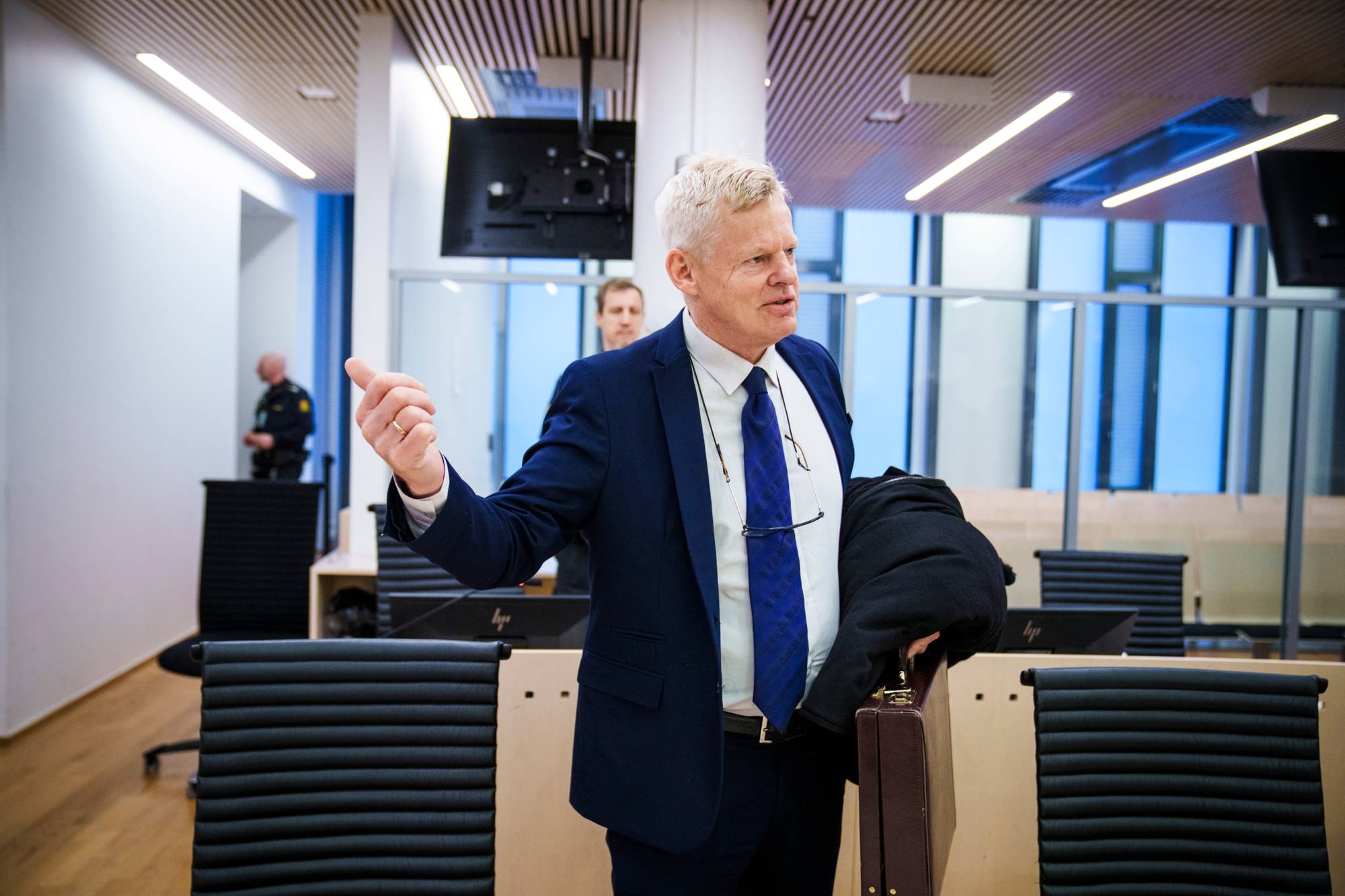 En mann i dress med slips gestikulerer. Han holder en kappe og en koffert. I bakgrunnen sitter en annen mann og en person i uniform. Det ser ut som et rettslokale.
