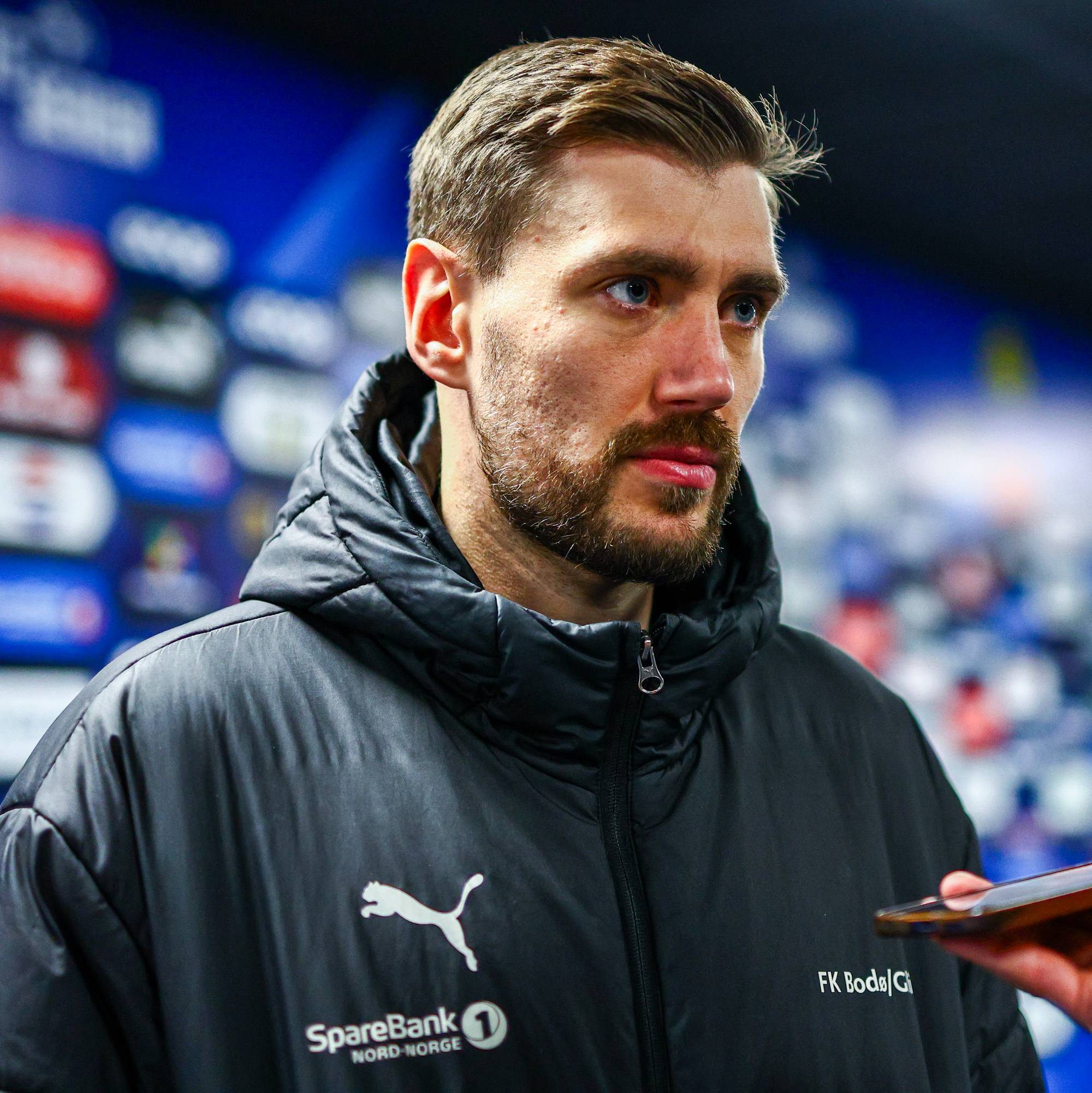 En mann med kort, brunt hår og skjegg intervjues foran en blå bakgrunn med sponsorlogoer. Mannen har på seg en mørk jakke med logoer for SpareBank 1 og FK Bodø/Glimt. En hånd holder en mobiltelefon opp mot ham, sannsynligvis for å ta opp intervjuet. Bakgrunnen viser et publikum med blå og hvite farger. (Bildebeskrivelsen er laget av en KI-tjeneste)