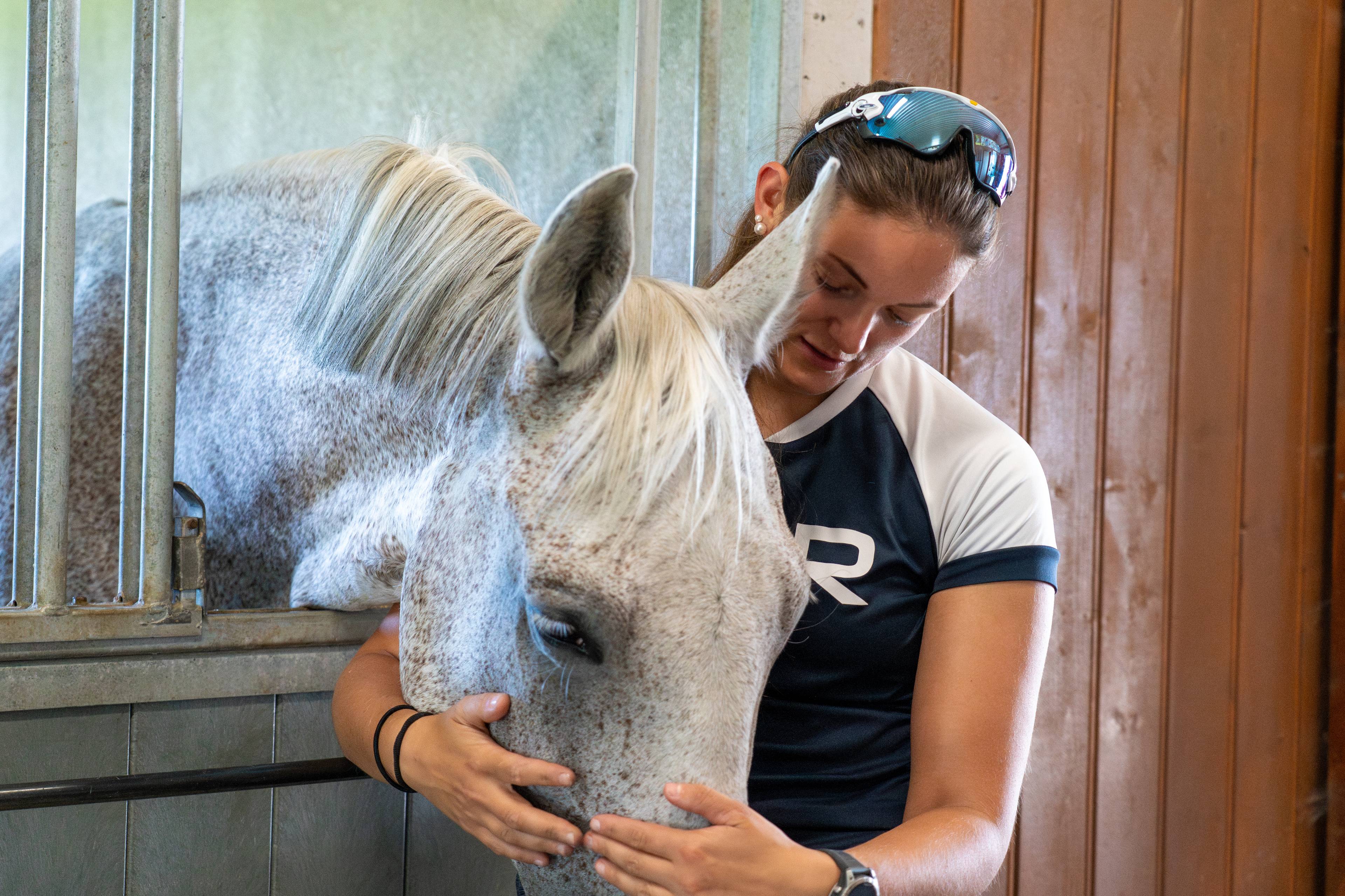 Kvinne i stall klemmer ein grå hest som stikker hovudet ut av ein boks. (Bildebeskrivelsen er laget av ein KI-teneste)