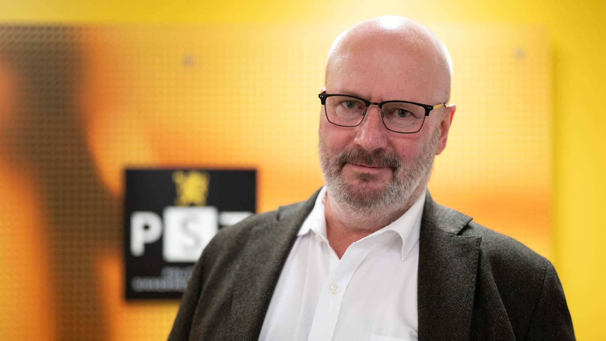 En mann i mørk jakke og hvit skjorte står foran en vegg med logoen til PST. Bakgrunnen er i gult og oransje, og gir kontekst til bildet. Mannen ser alvorlig ut, noe som kan indikere en viktig situasjon. Ingen andre personer er synlige i bildet. (Bildebeskrivelsen er laget av en KI-tjeneste)