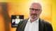 Tom Balgaard / NRK En mann i mørk jakke og hvit skjorte står foran en vegg med logoen til PST. Bakgrunnen er i gult og oransje, og gir kontekst til bildet. Mannen ser alvorlig ut, noe som kan indikere en viktig situasjon. Ingen andre personer er synlige i bildet. (Bildebeskrivelsen er laget av en KI-tjeneste)