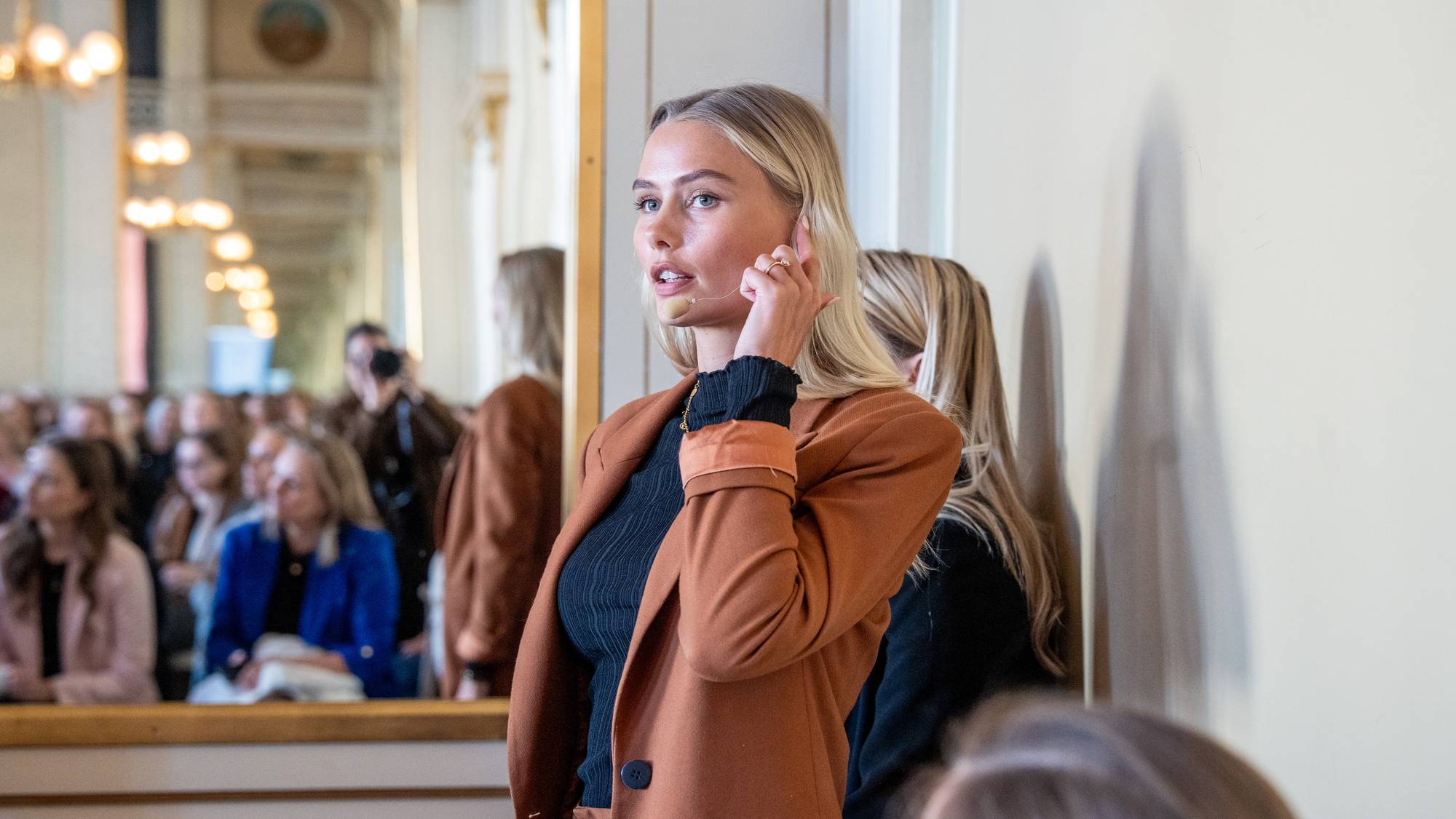En ung kvinne med blondt hår snakker til et publikum. Hun har på seg en brun blazer over en svart genser. I bakgrunnen ser man et publikum reflektert i et stort speil. Noen i publikum holder opp kameraer for å ta bilder. (Bildebeskrivelsen er laget av en KI-tjeneste)