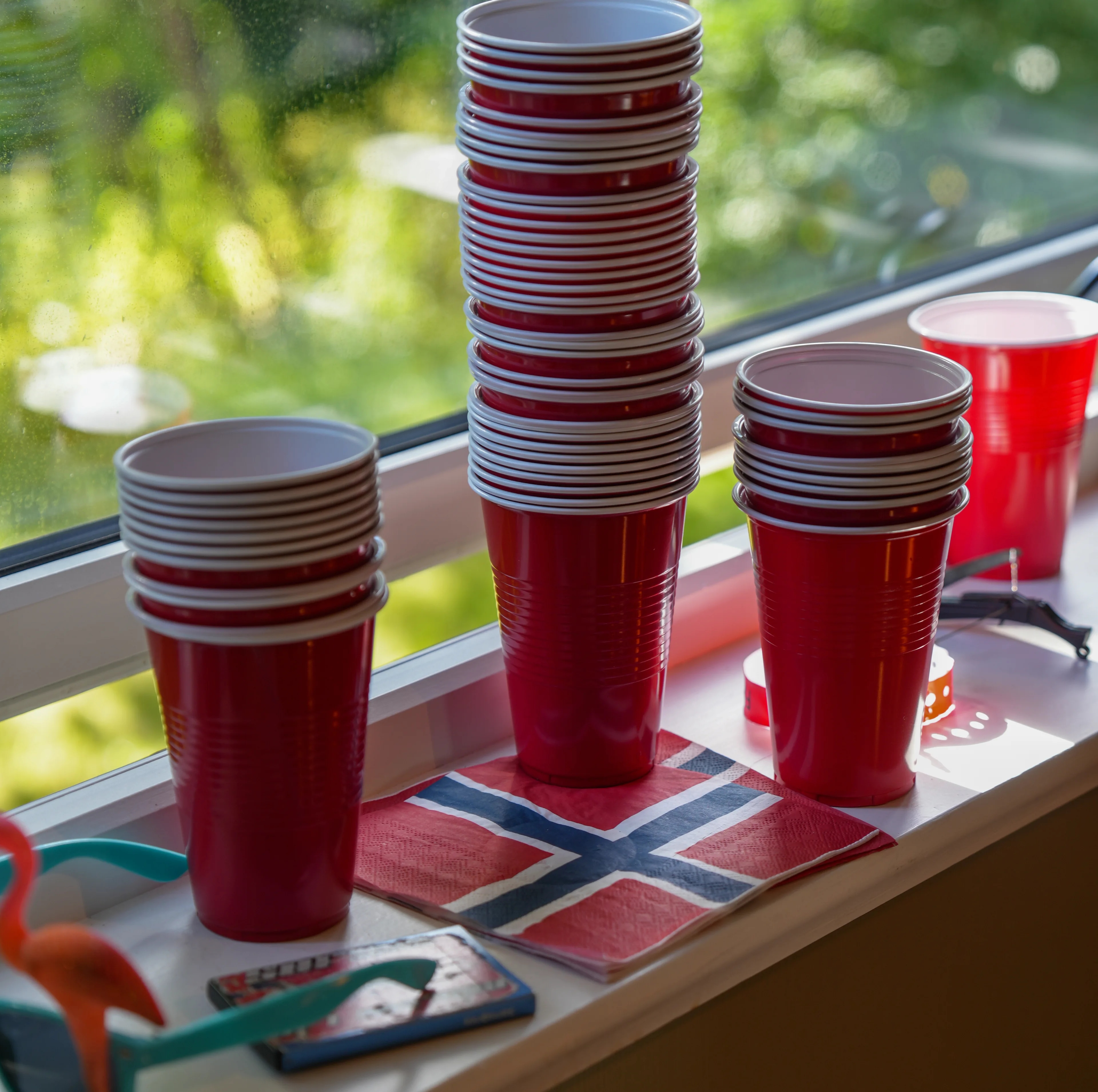 Stabler med røde plastkopper og en serviett med det norske flagget står i en vinduskarm. (Bildebeskrivelsen er laget av en KI-tjeneste)